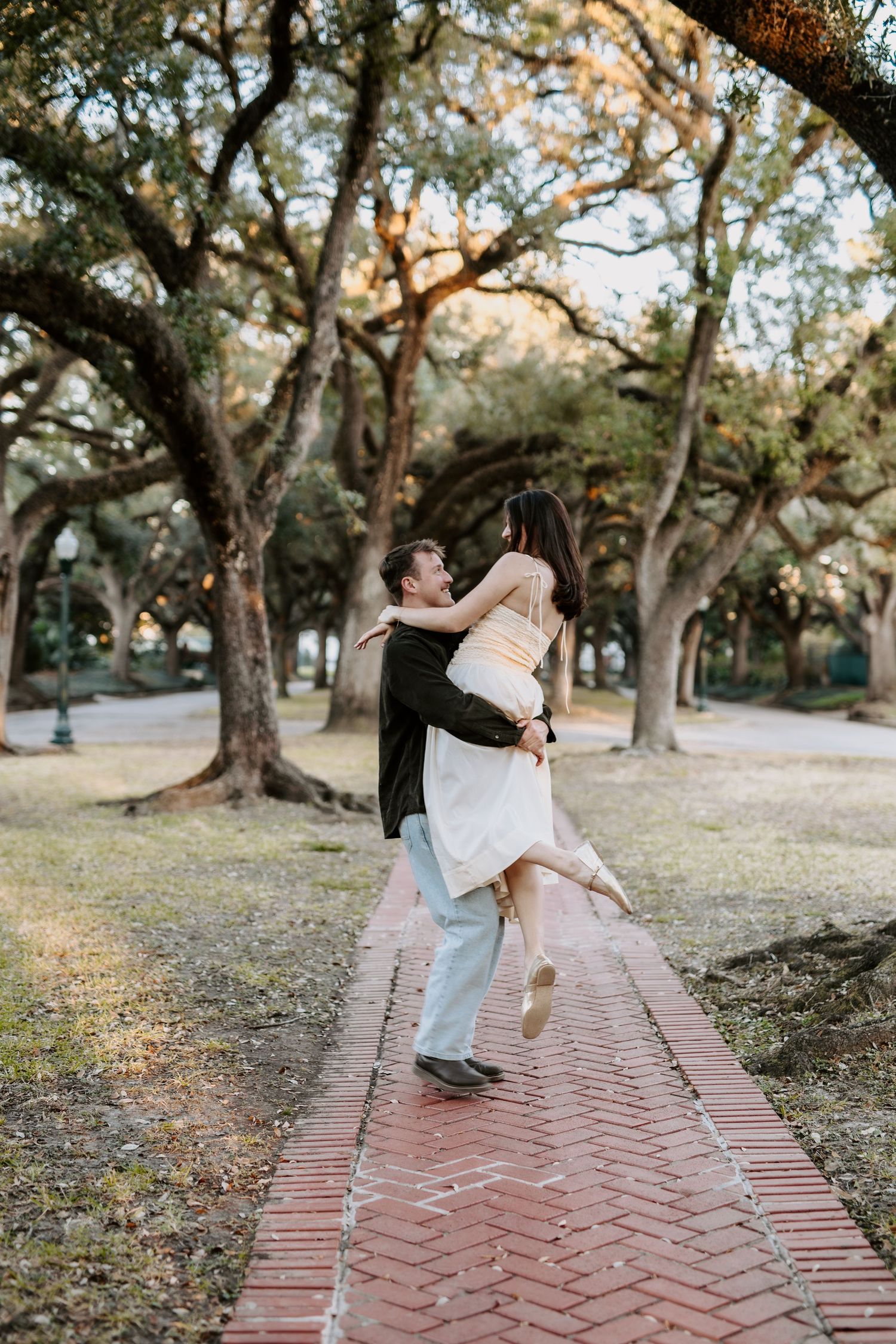 Groom lifting bride on brick pathway surrounded by large oak trees during golden hour engagement session.