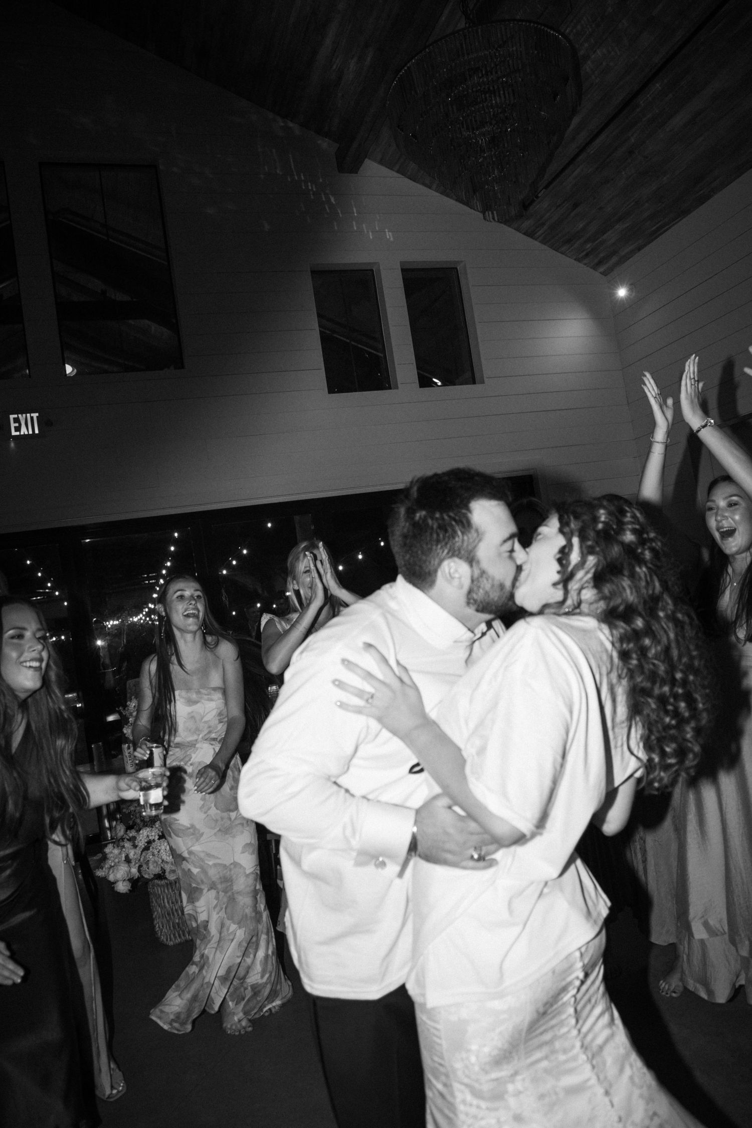 A newlywed couple shares a passionate kiss on the dance floor surrounded by cheering wedding guests.