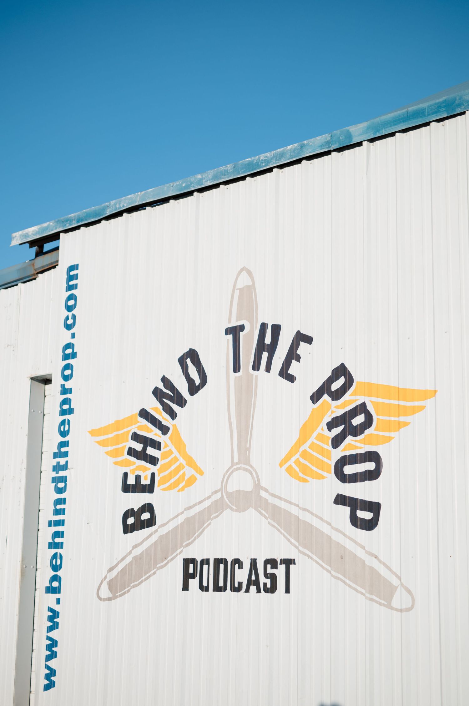 Behind the Prop aviation podcast logo painted on a white metal hangar wall under a clear blue sky.