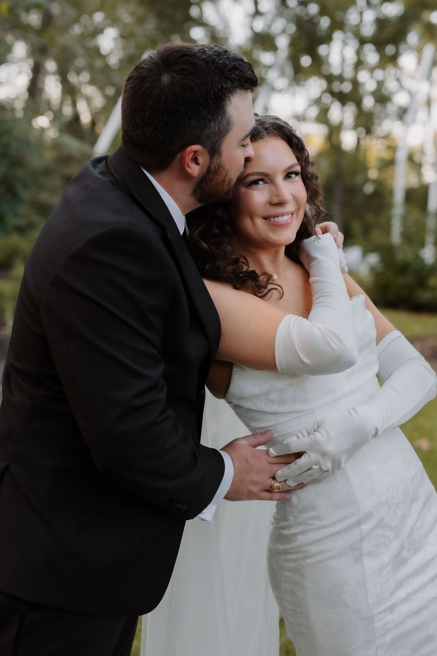 Groom in black suit embraces smiling bride in white gown and gloves in outdoor setting.