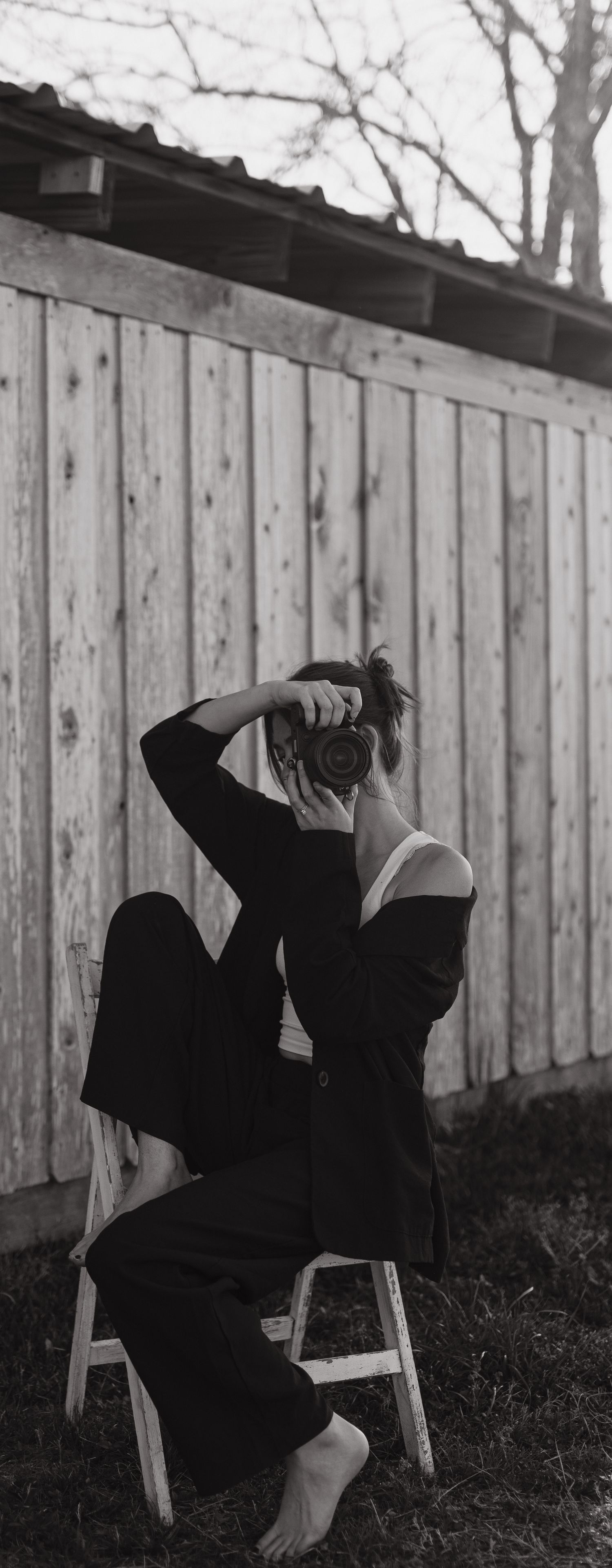 Black and white photo of barefoot woman in black outfit sitting on stool photographing outdoors.