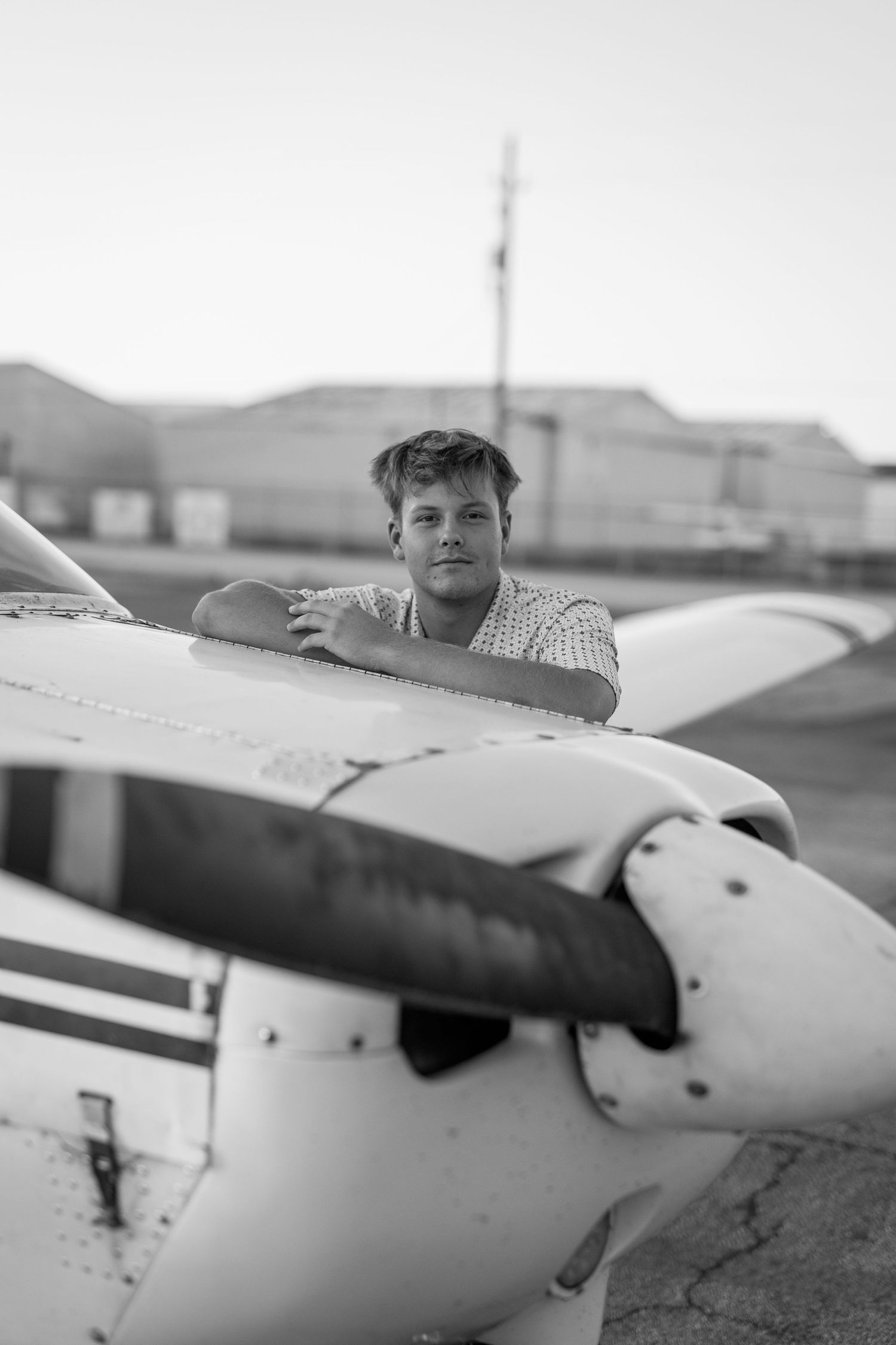 Young man in sunglasses leaning over white propeller plane wing at airfield in black and white.