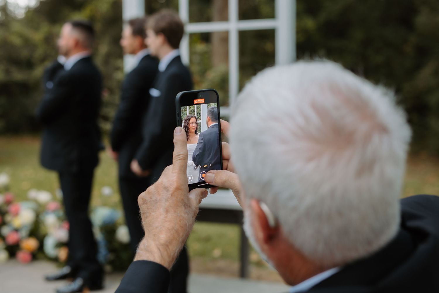 Guest photographs bride and groom on smartphone during outdoor wedding ceremony with groomsmen in background.