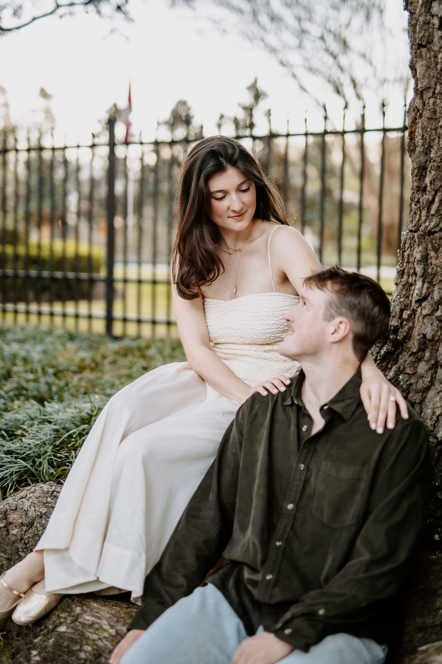 Engaged couple sitting by a tree near an iron fence in a park setting.