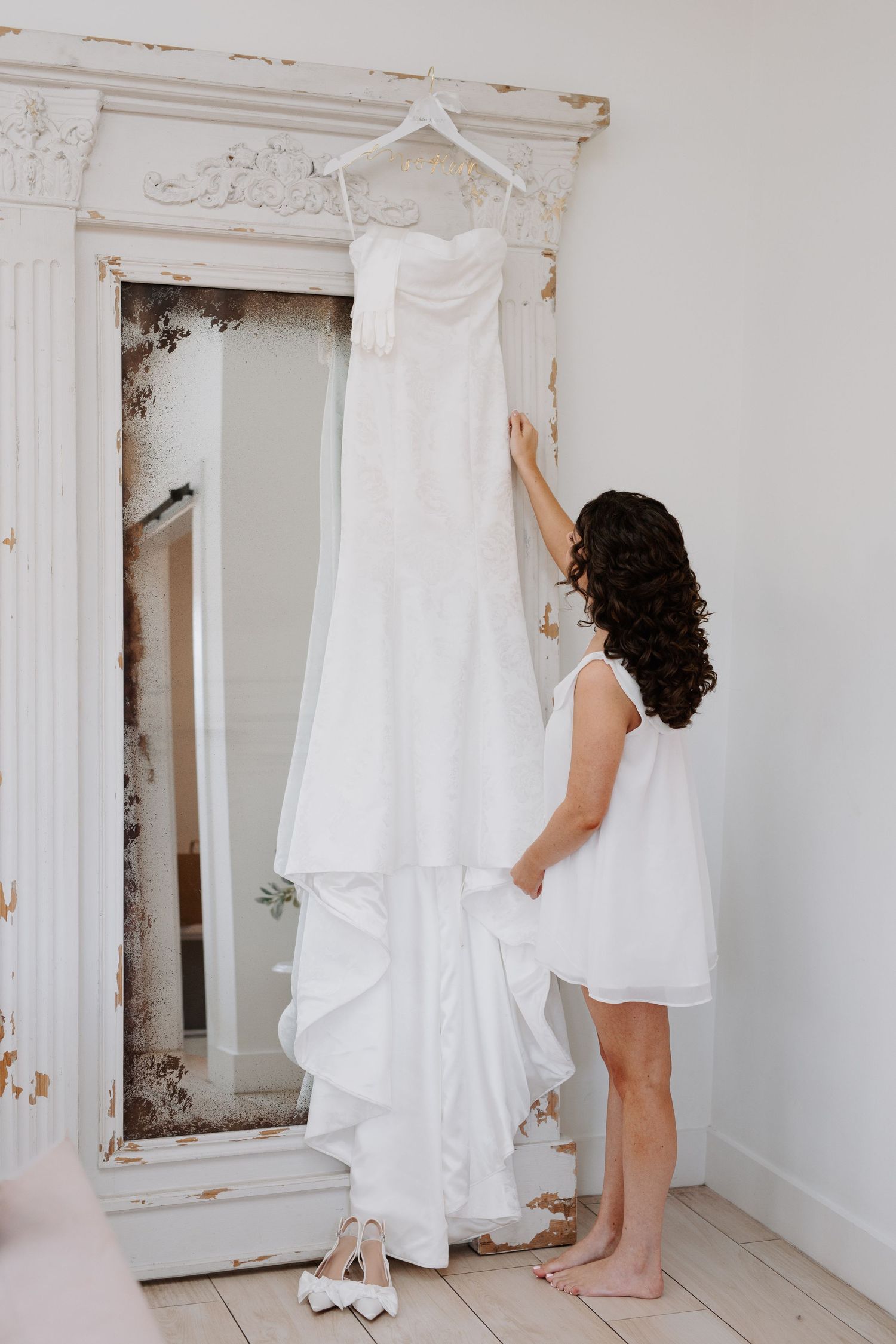 Bride in white dress reaching up to touch her wedding gown hanging on an ornate vintage mirror.