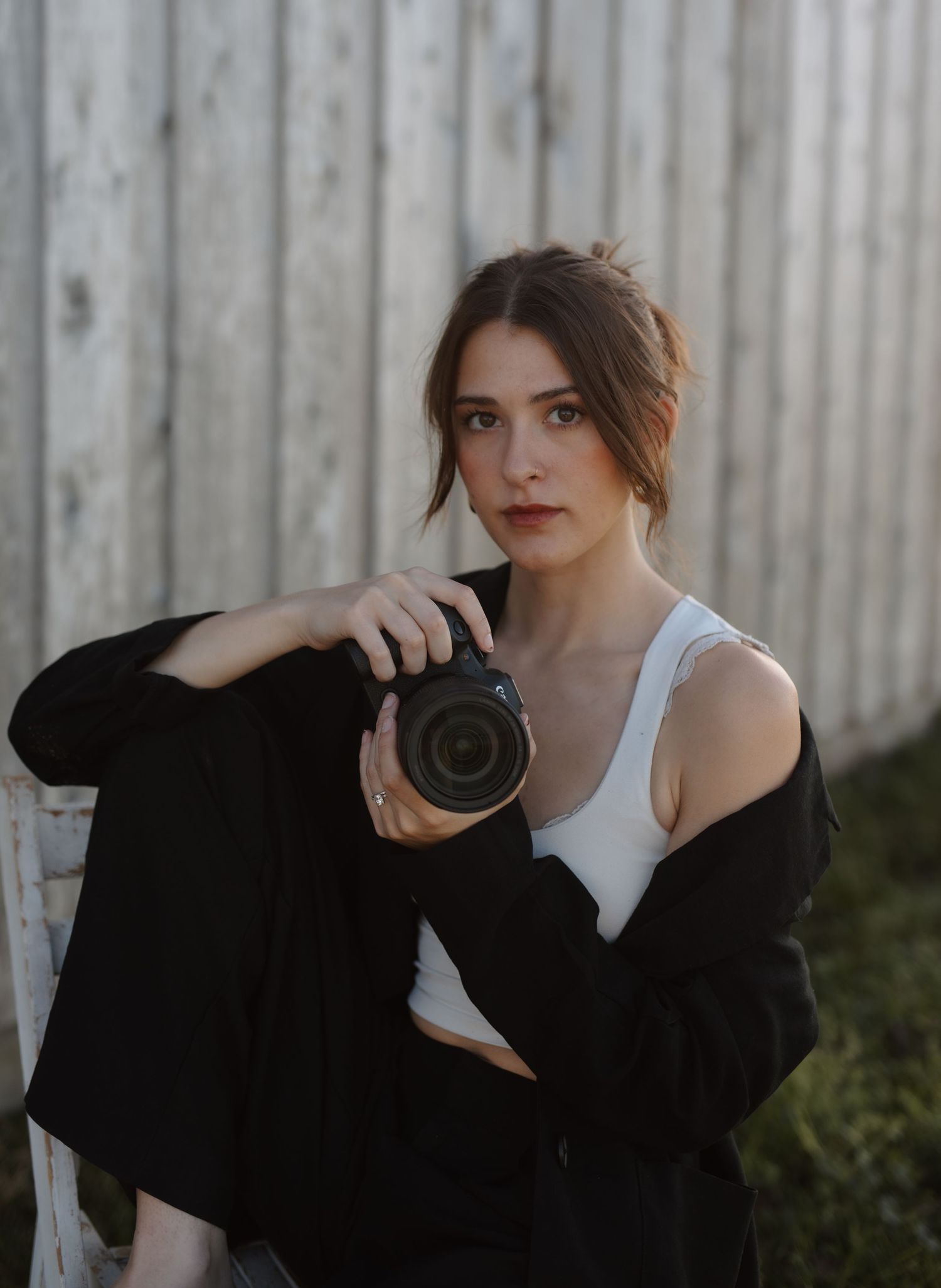 Young woman in black blazer and white top posing with DSLR camera seated on white chair outdoors.