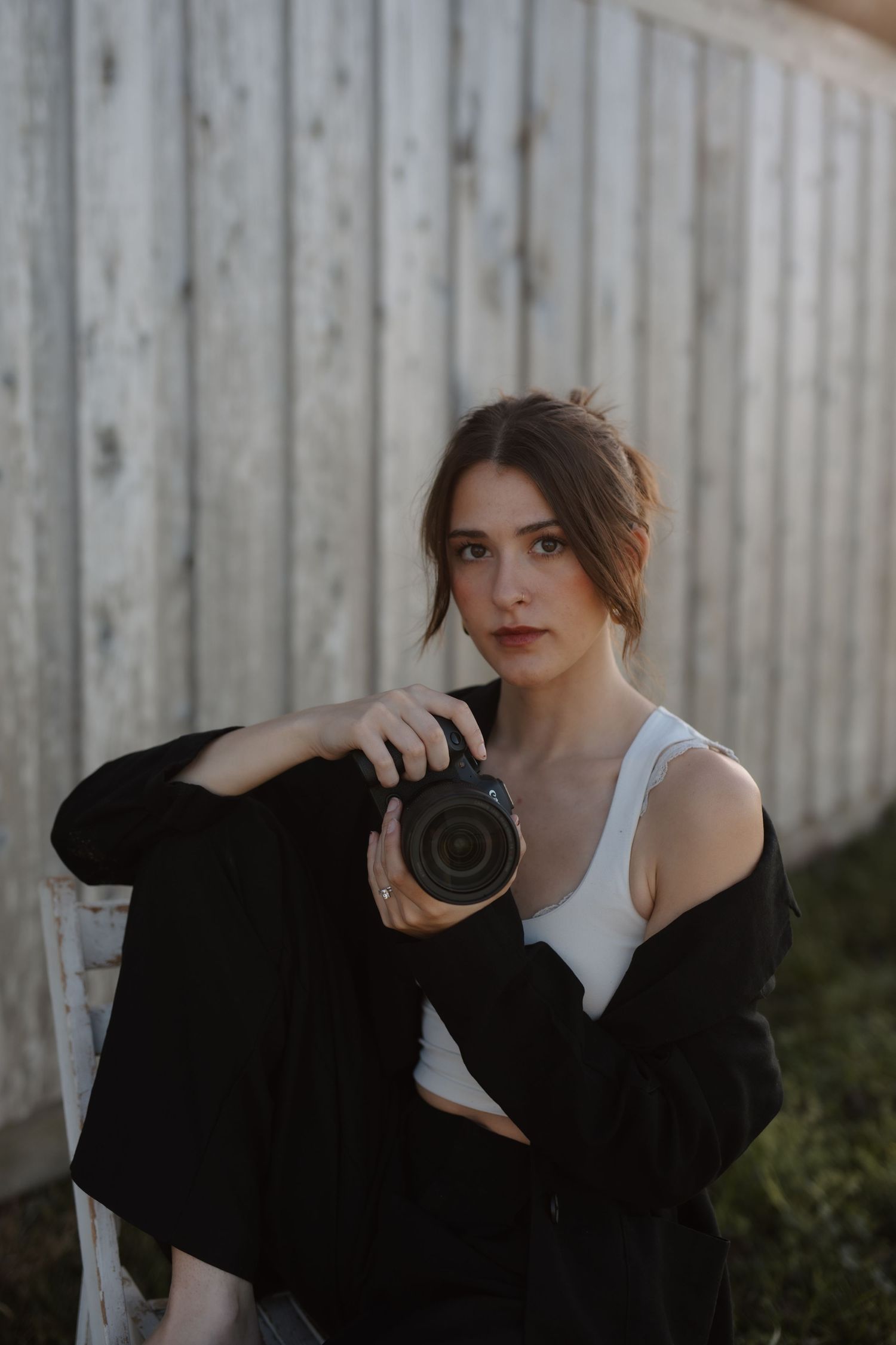 Young female photographer in black blazer holding camera while seated against wooden fence.