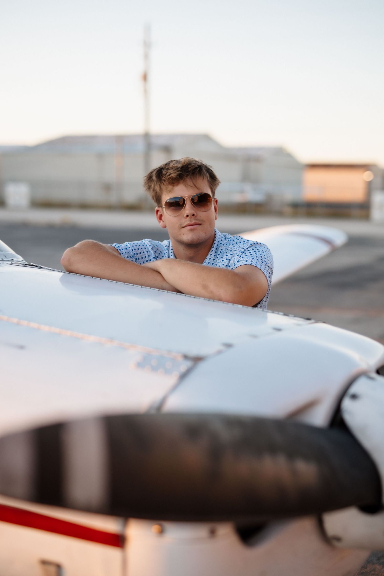 Young man in sunglasses resting on white small plane wing at sunset airfield.