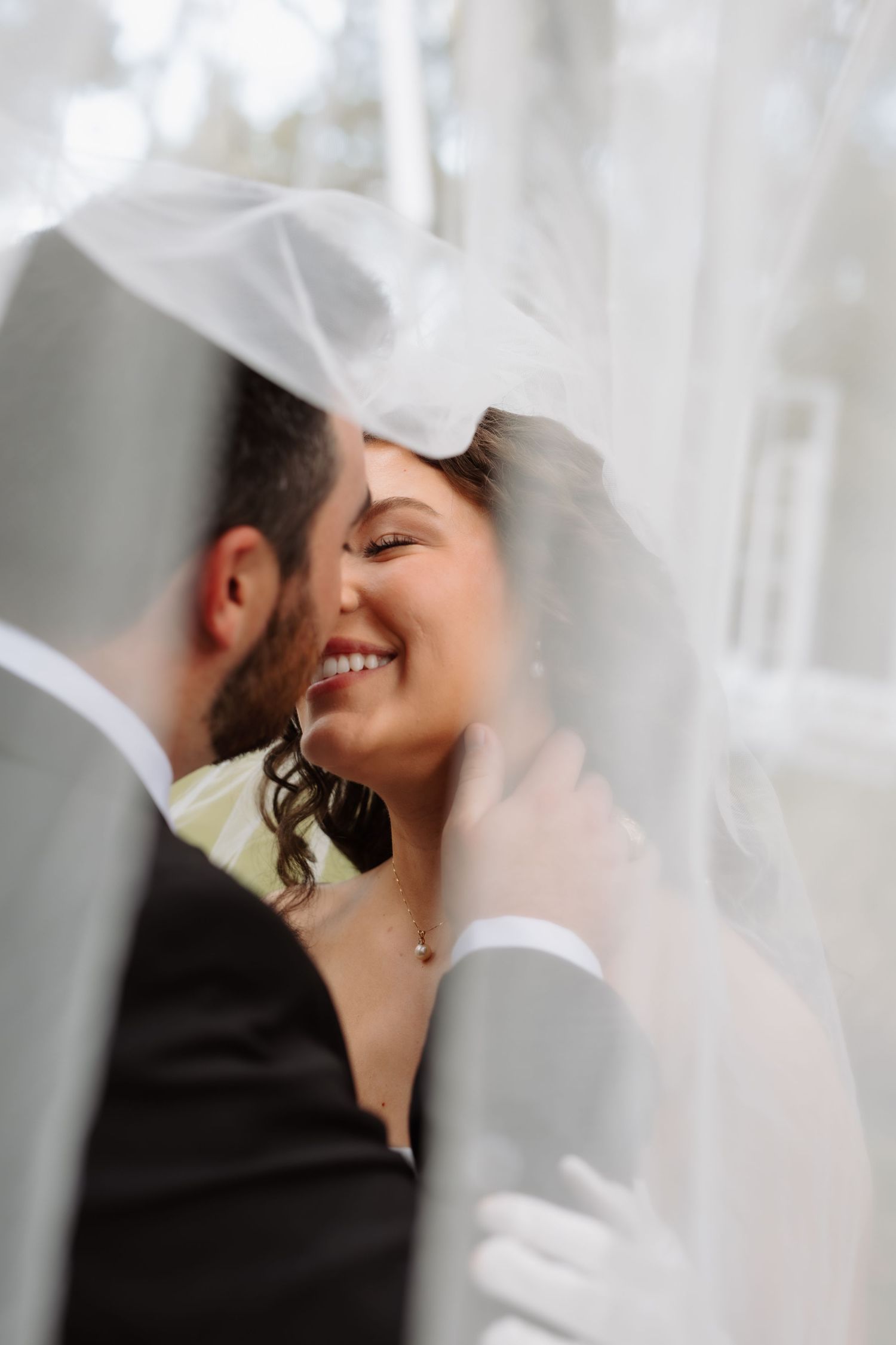 Bride smiles joyfully at groom under flowing white veil in intimate outdoor wedding portrait.