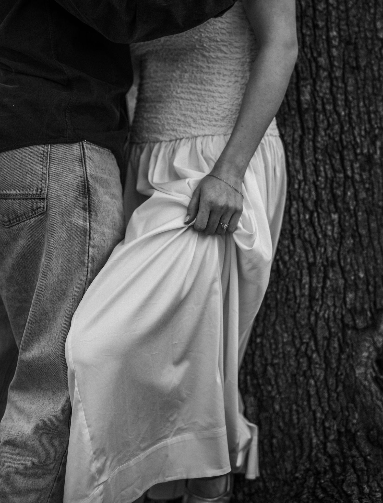 Black and white photo of couple standing by tree, groom holding brides white dress skirt gently.