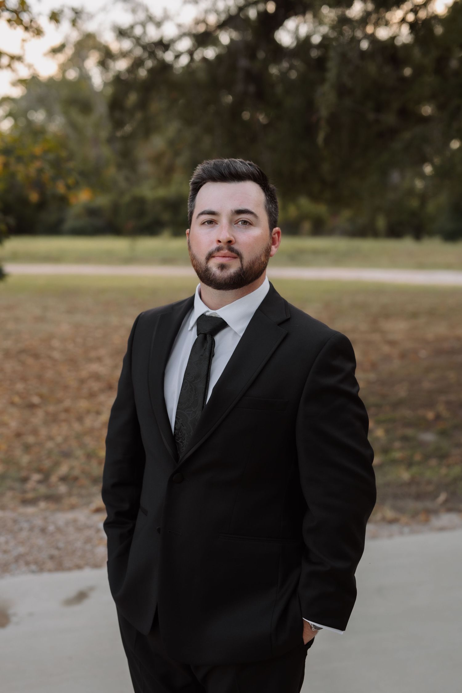 Groom in black suit and tie standing outdoors at wedding venue with trees in background.
