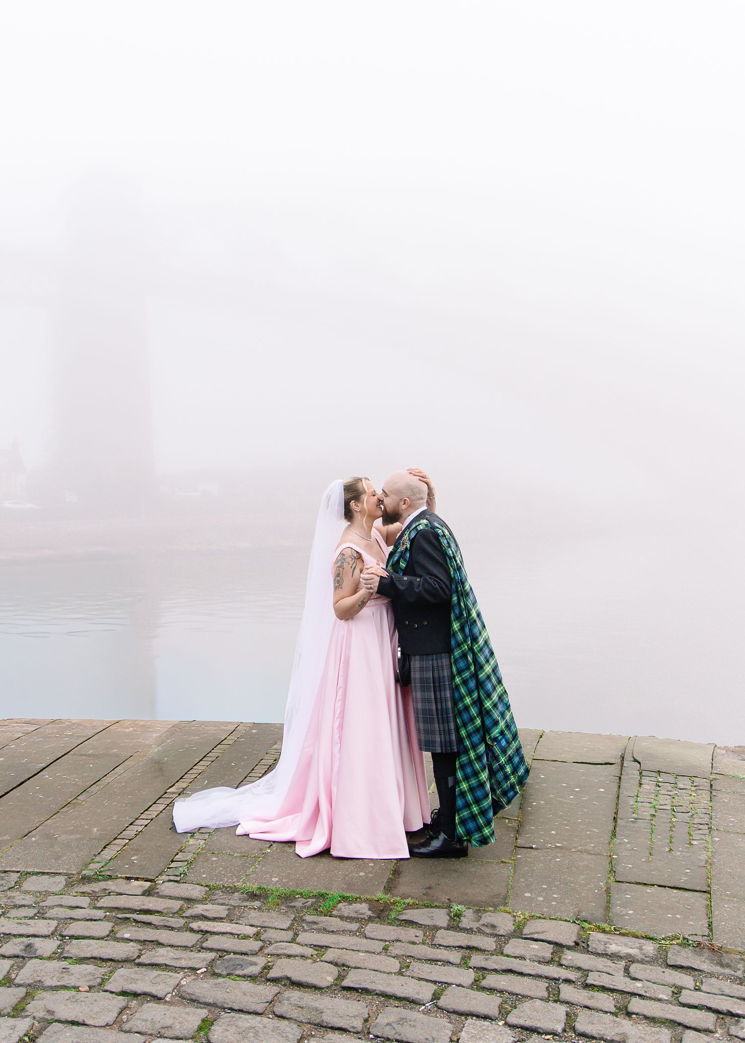 Couple in wedding attire sits on cobblestone steps in soft lighting.