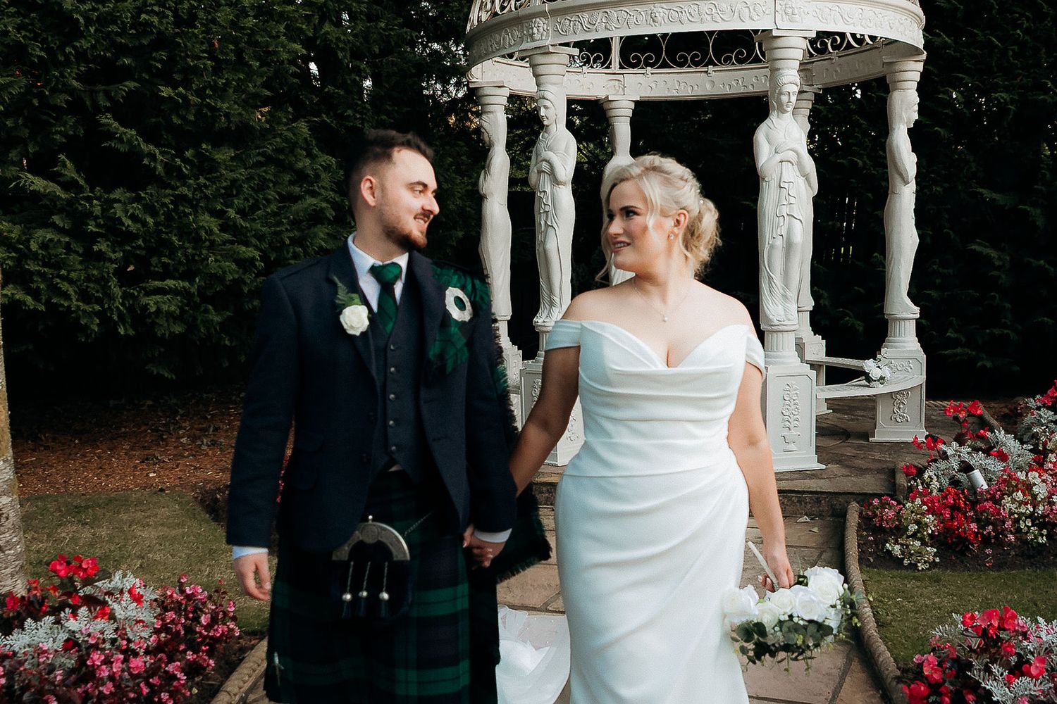 Bride in white dress and groom in kilt standing by garden gazebo with red flowers.