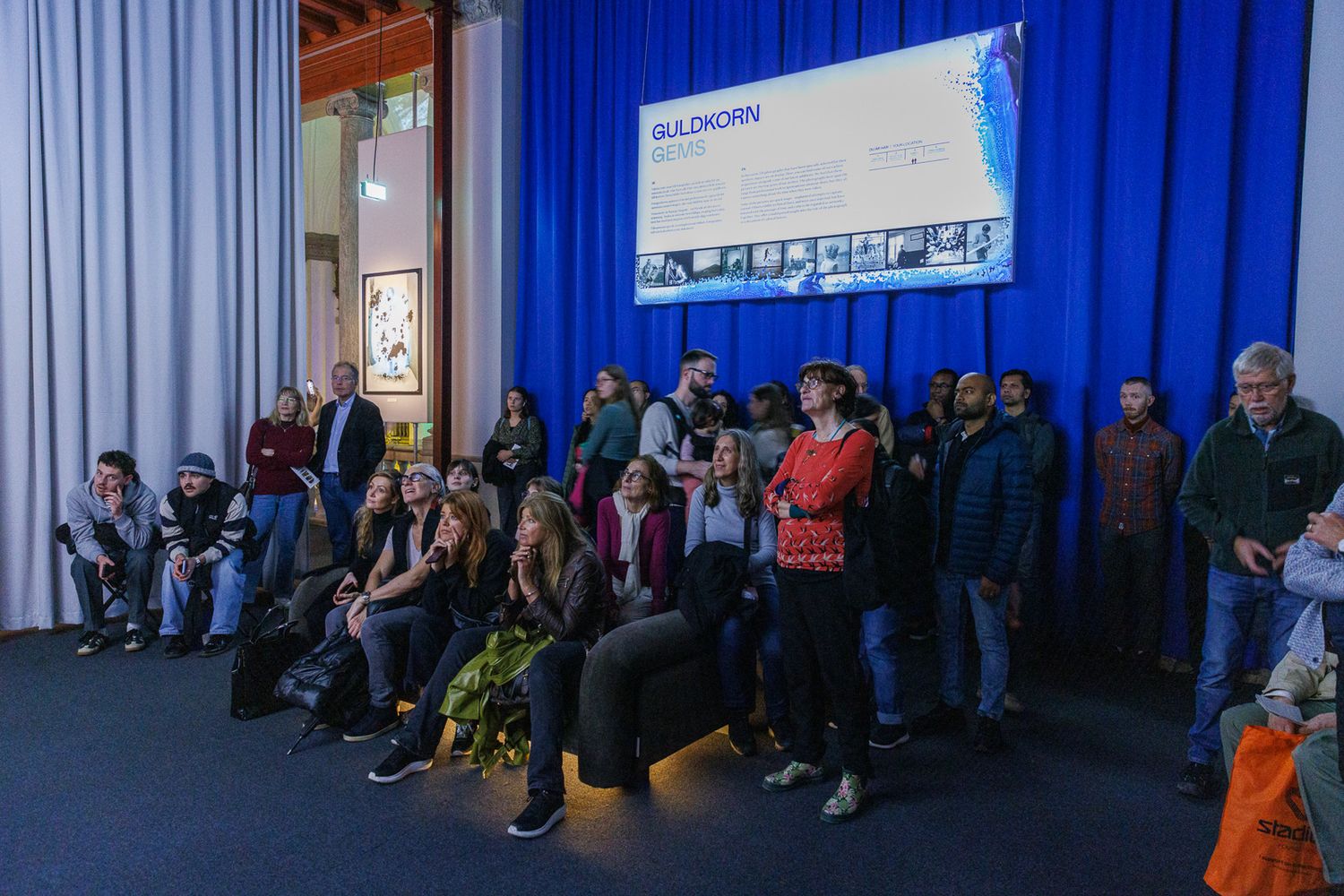 Audience members seated along a wall with blue curtains in an auditorium setting.
