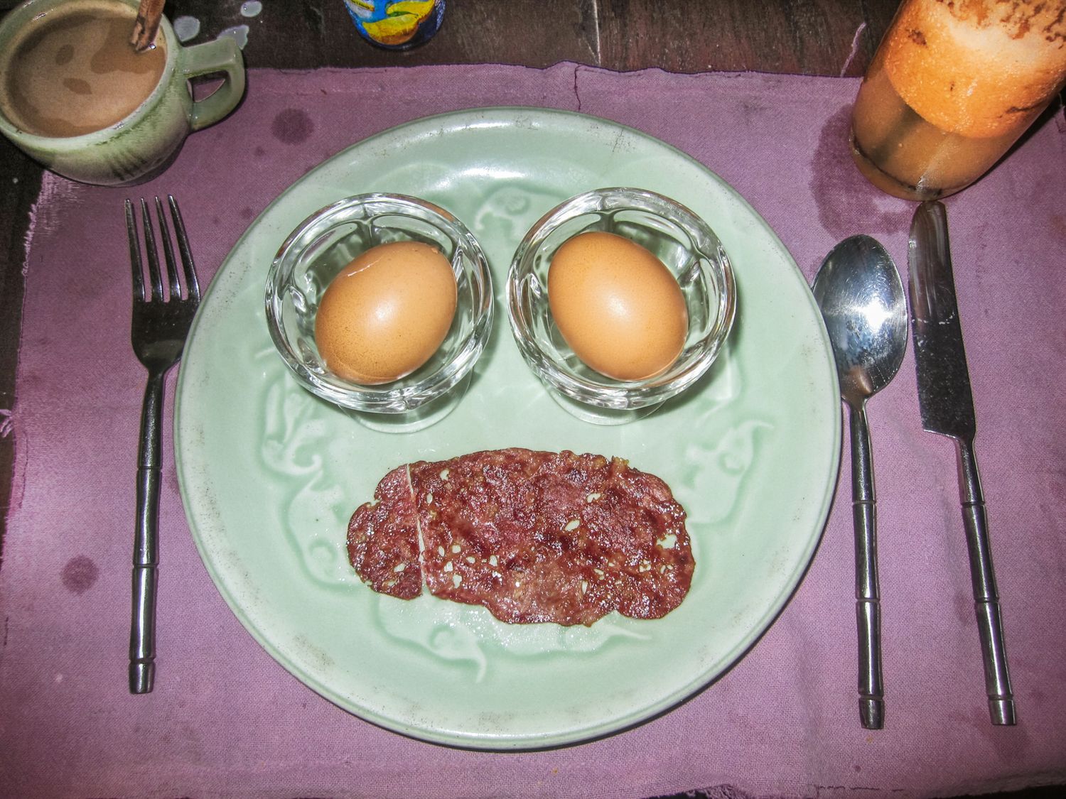 A plate with two eggs in glass holders and sliced corned beef, flanked by a fork and spoon, with drinks nearby.