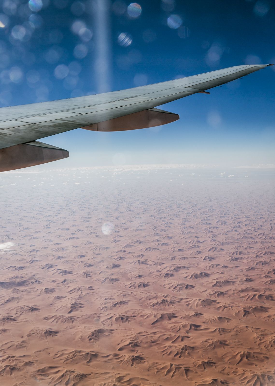 Airplane wing view over vast desert landscape with rippling sand dunes below.