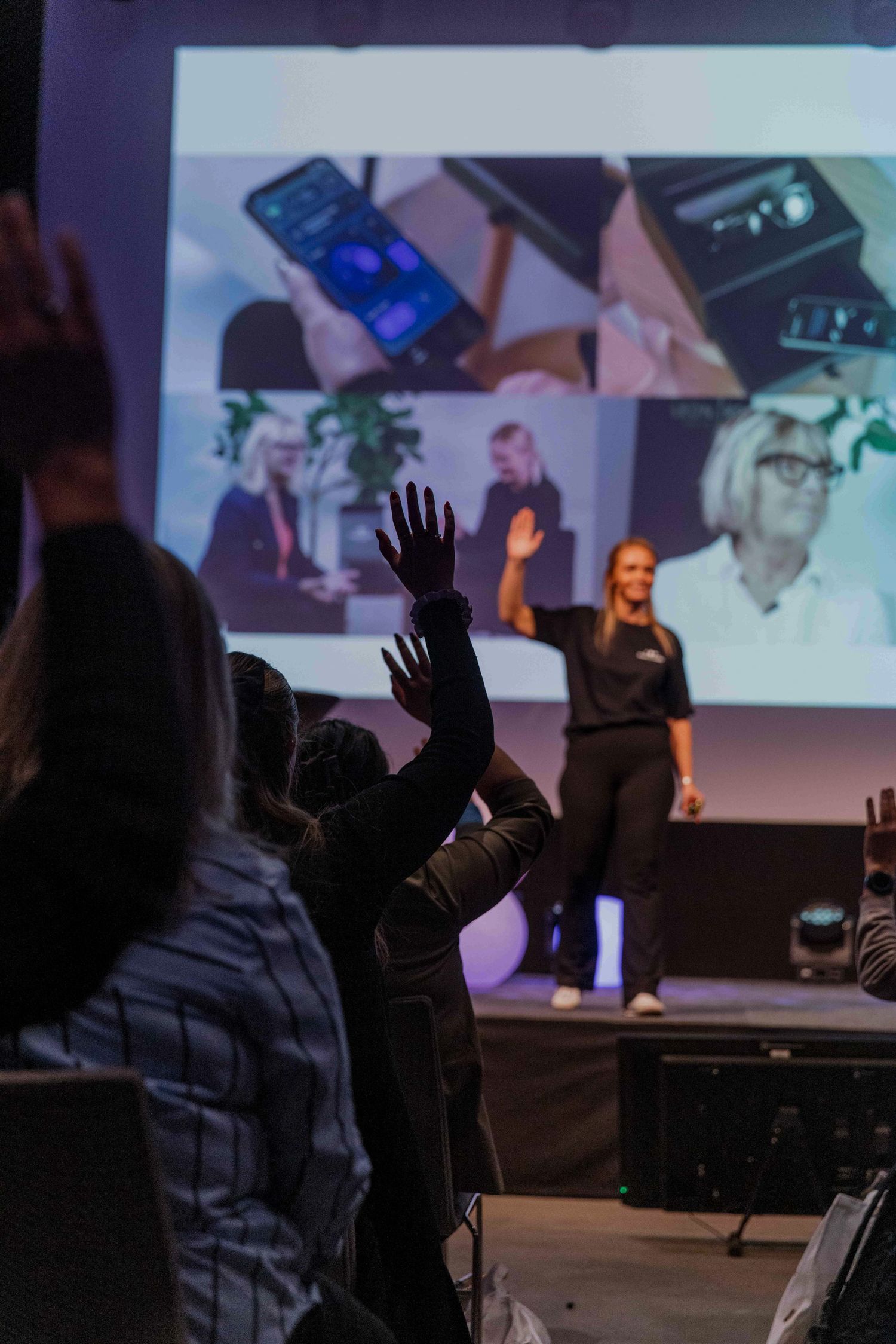 Audience members raising hands during a presentation with projected screens in background.