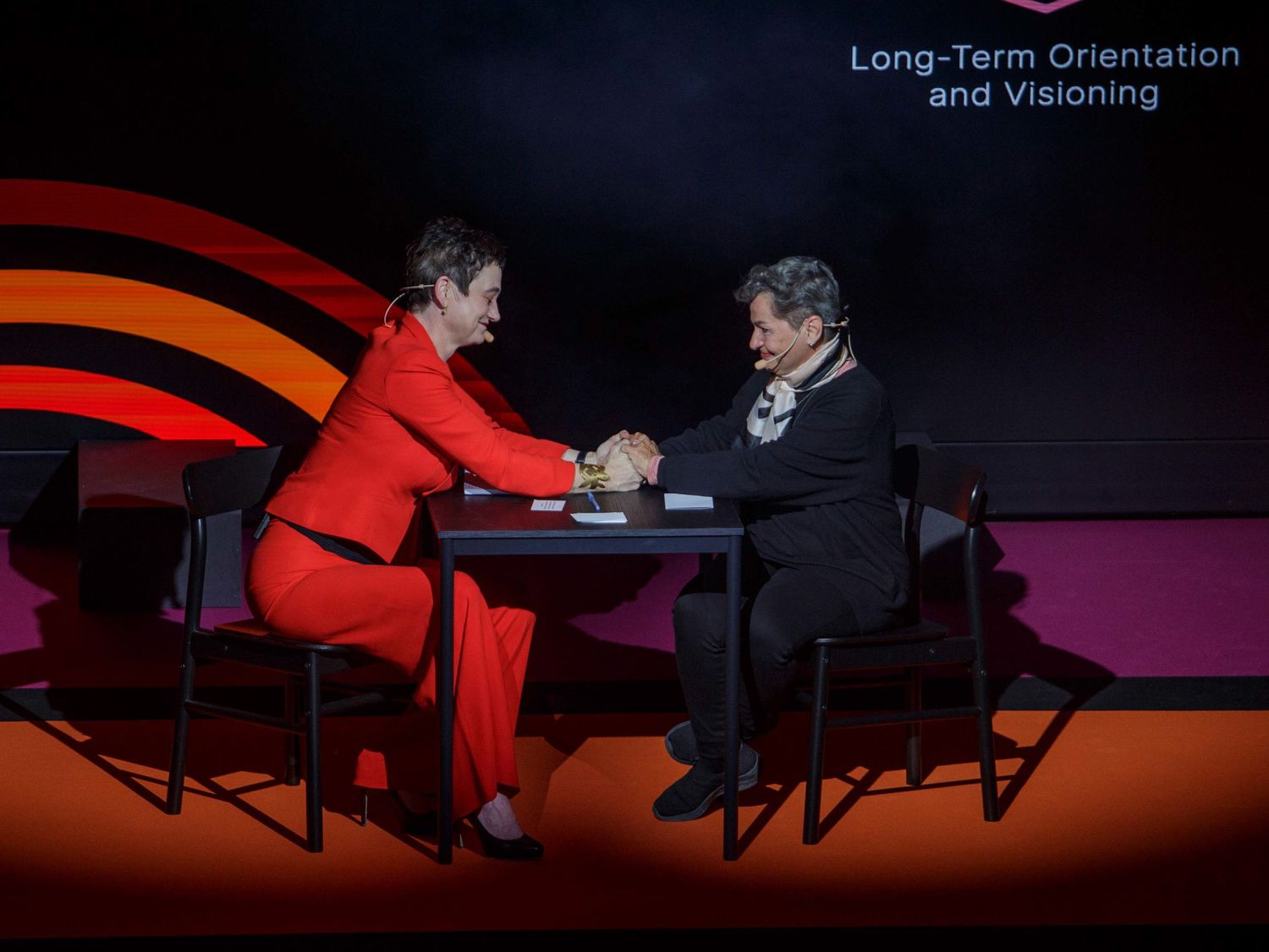 Two speakers engage in a discussion on stage at a conference with dramatic red lighting.