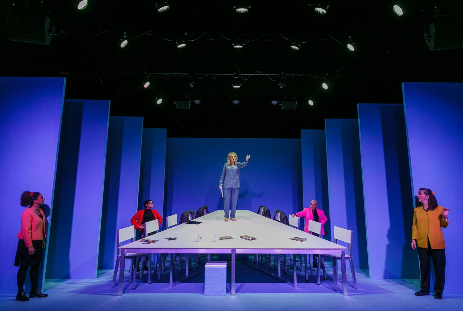 Wide shot of a minimalist stage set with white chairs against deep blue lighting.