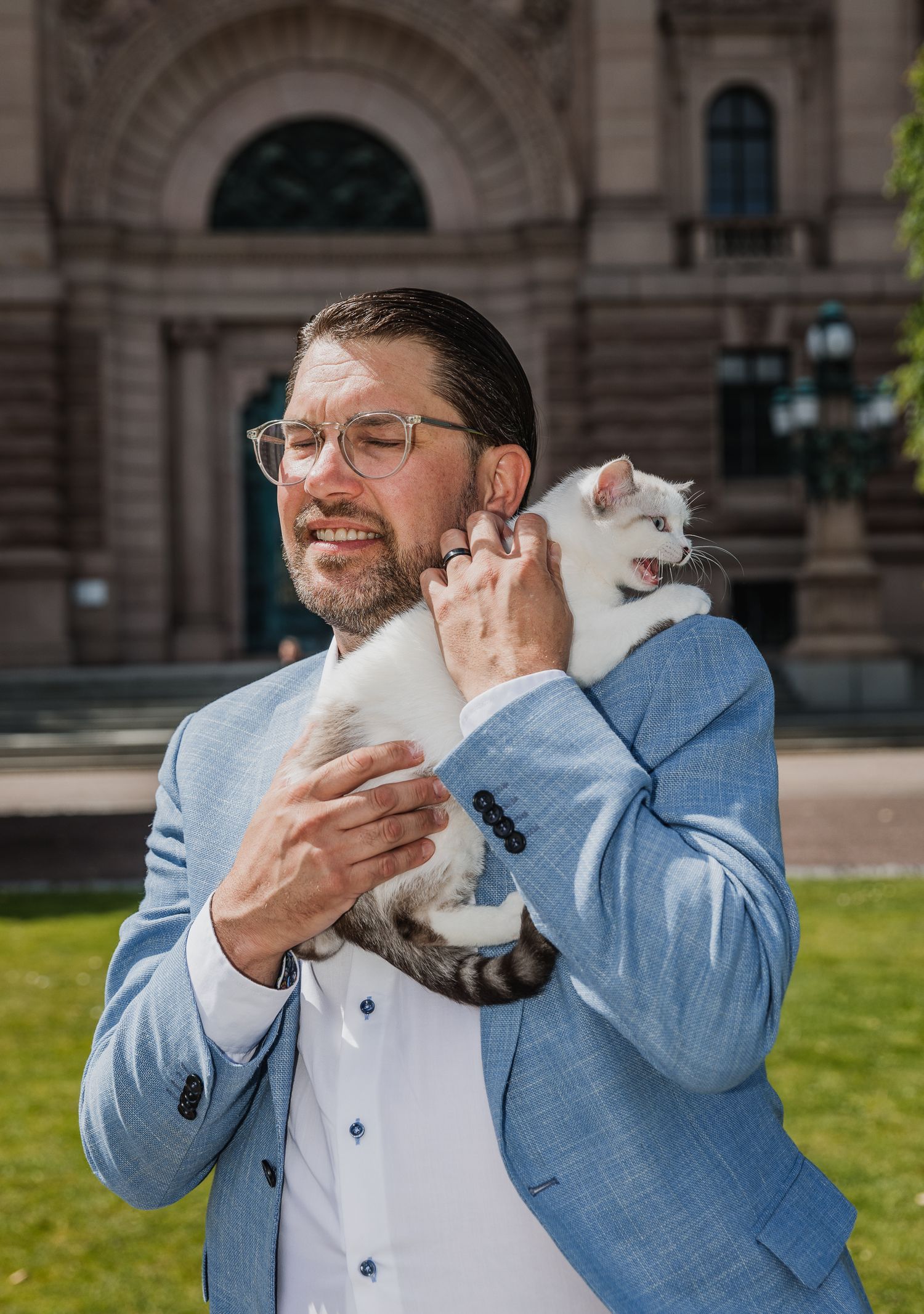 Man in blue blazer holding a small white cat outside a historic building on a sunny day.