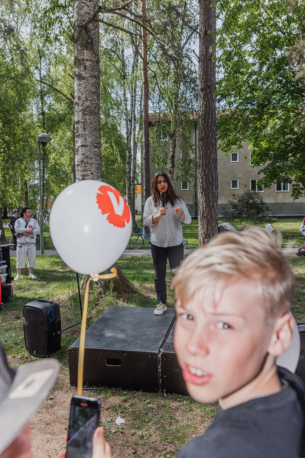 A blonde boy in foreground at outdoor event with speaker on stage near white balloon with red bird logo in sunny park.