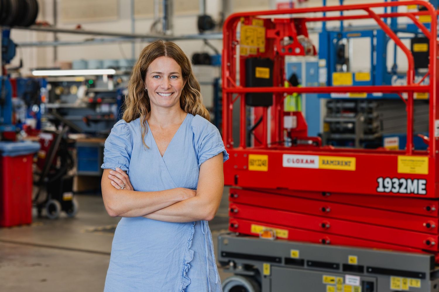 Woman smiling with arms crossed in front of a red Genie 1932ME scissor lift in an industrial warehouse.