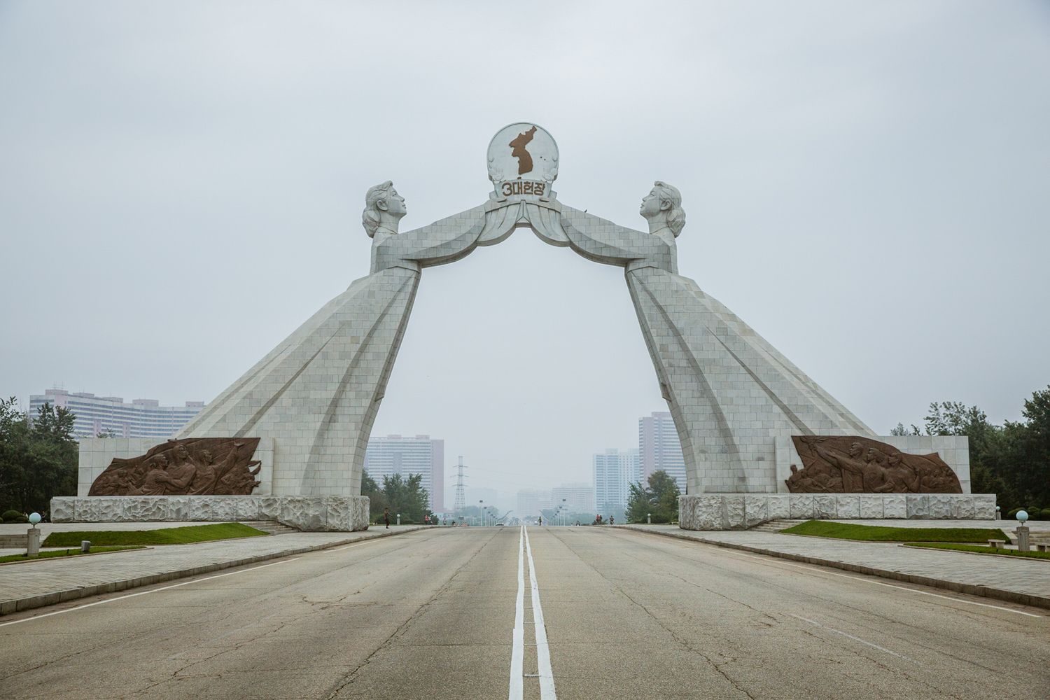 Monumental white concrete arch structure spans across a wide road in dramatic perspective.