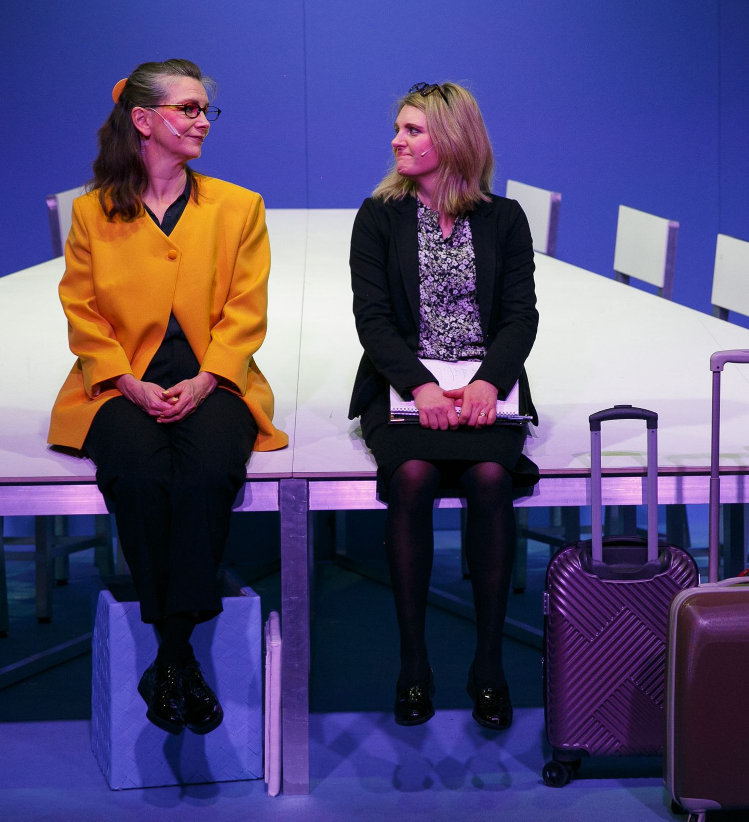 Performers seated on chairs against a purple-blue backdrop during a stage production.