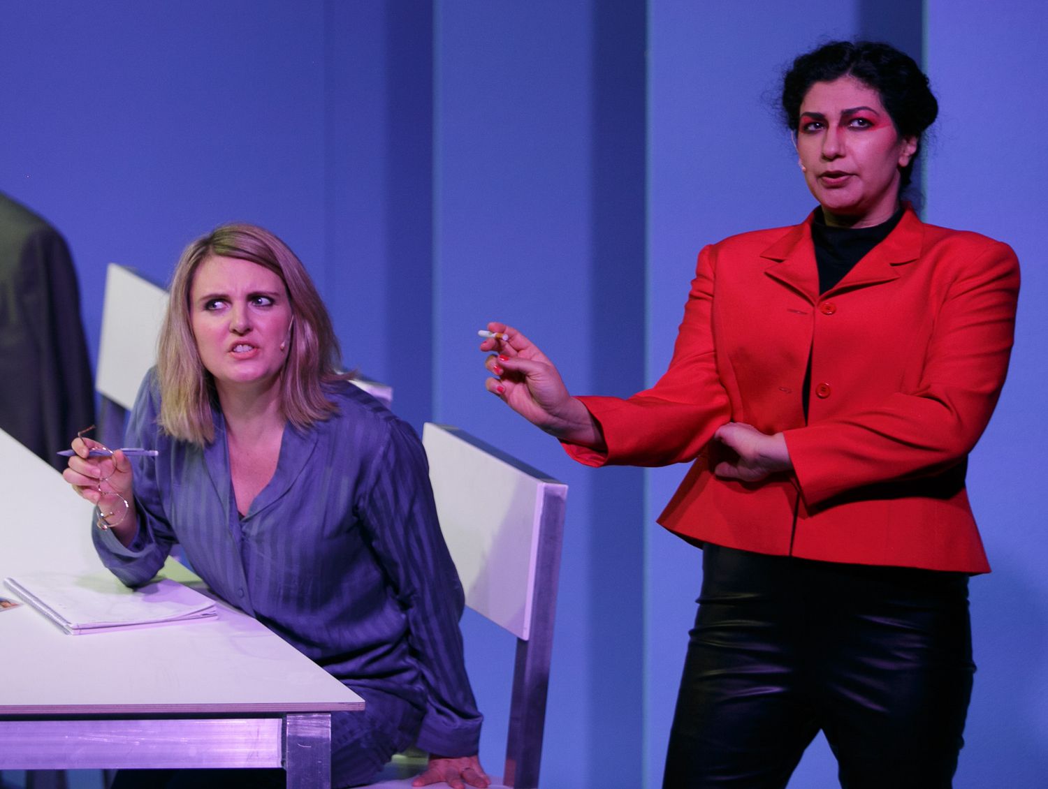 Performers in bright colored outfits gesture and interact during a theatrical scene on stage.