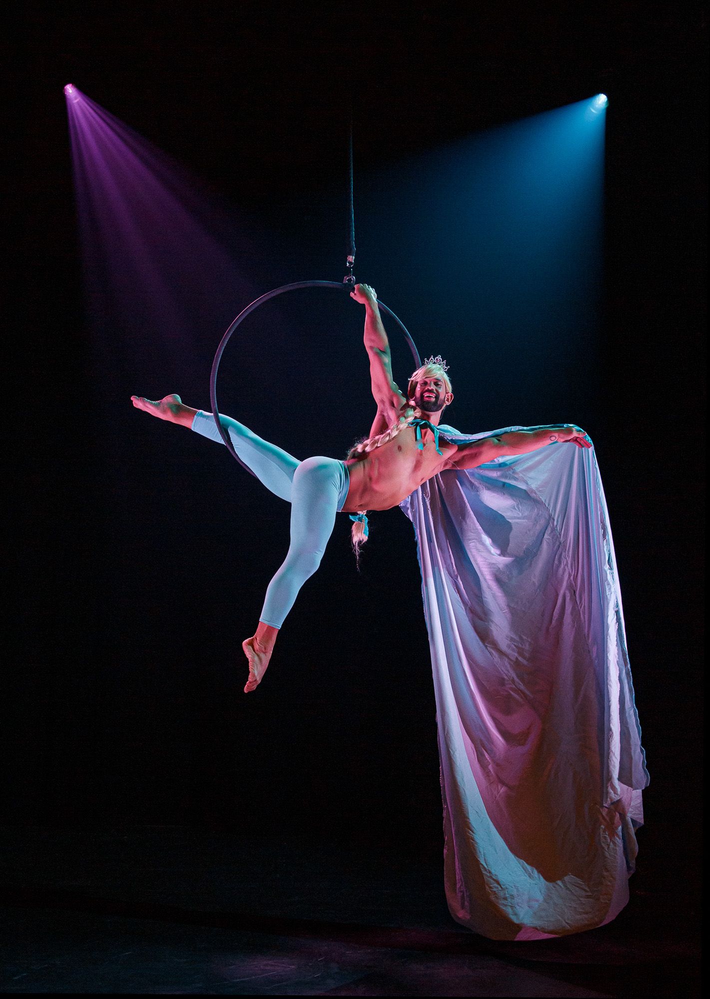 Aerial performer in flowing costume suspended on hoop under purple and blue spotlights.