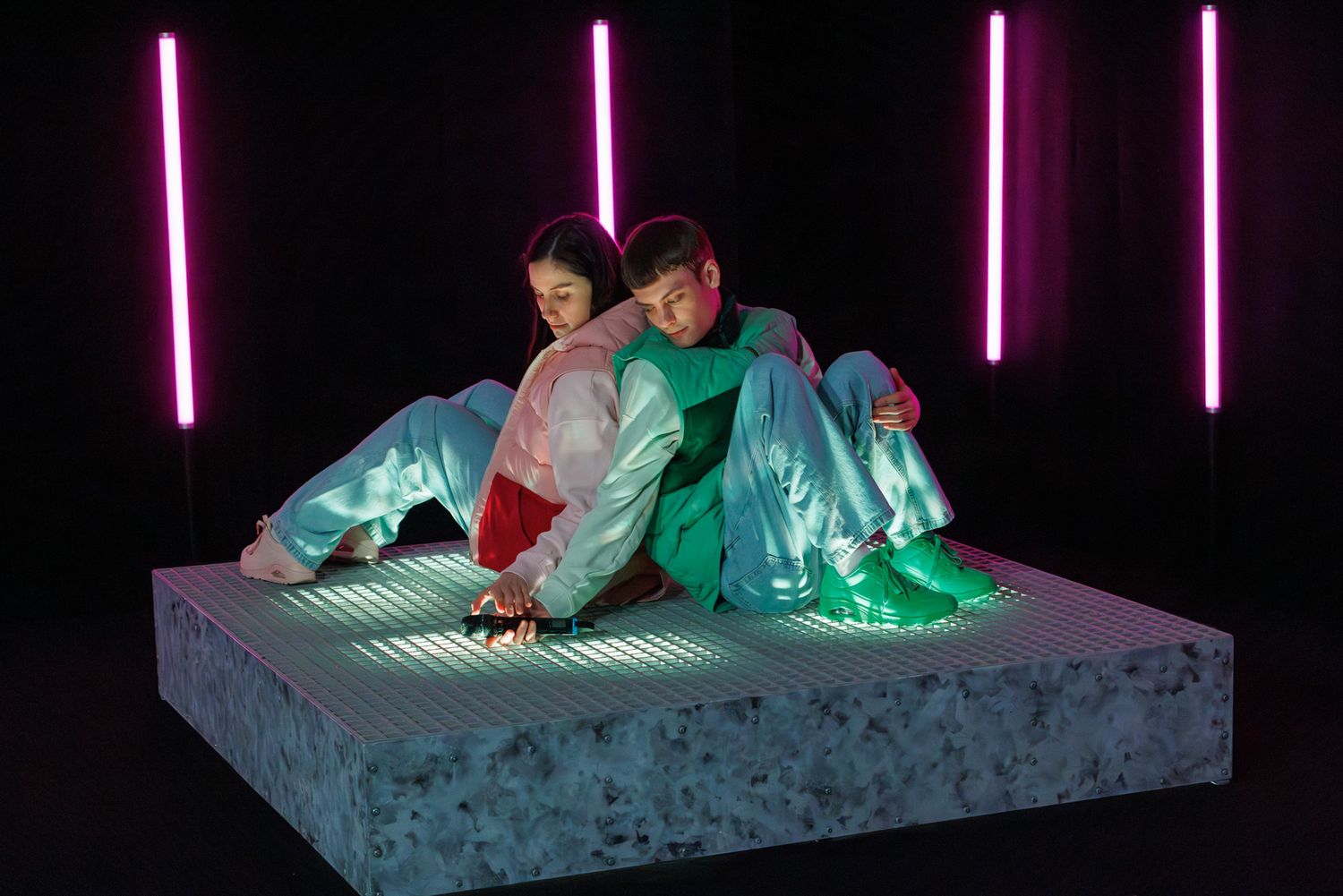 Two performers in colorful outfits posing on textured platform with pink neon lights.