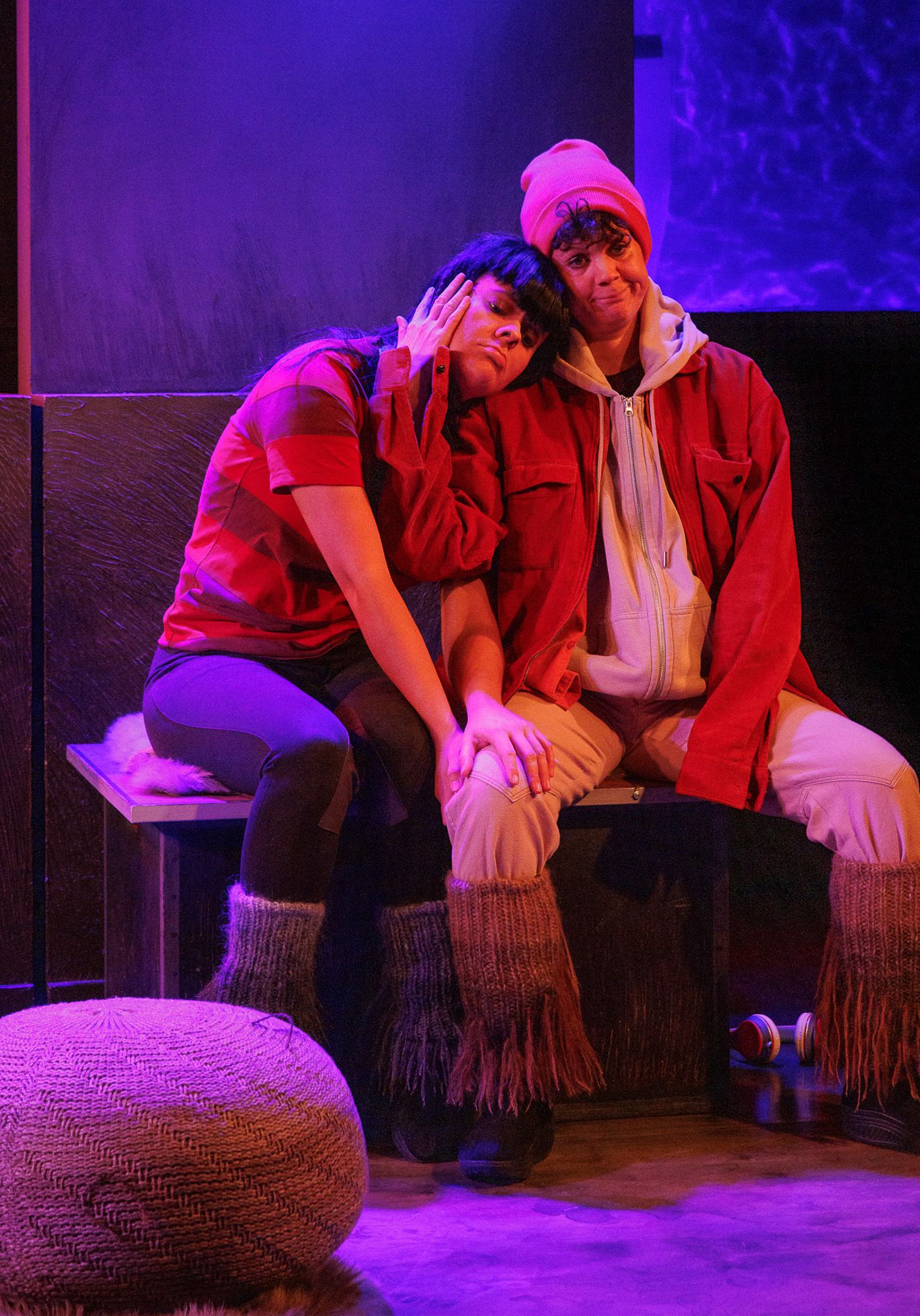 Theater scene with performers on textured ottomans under purple stage lighting.
