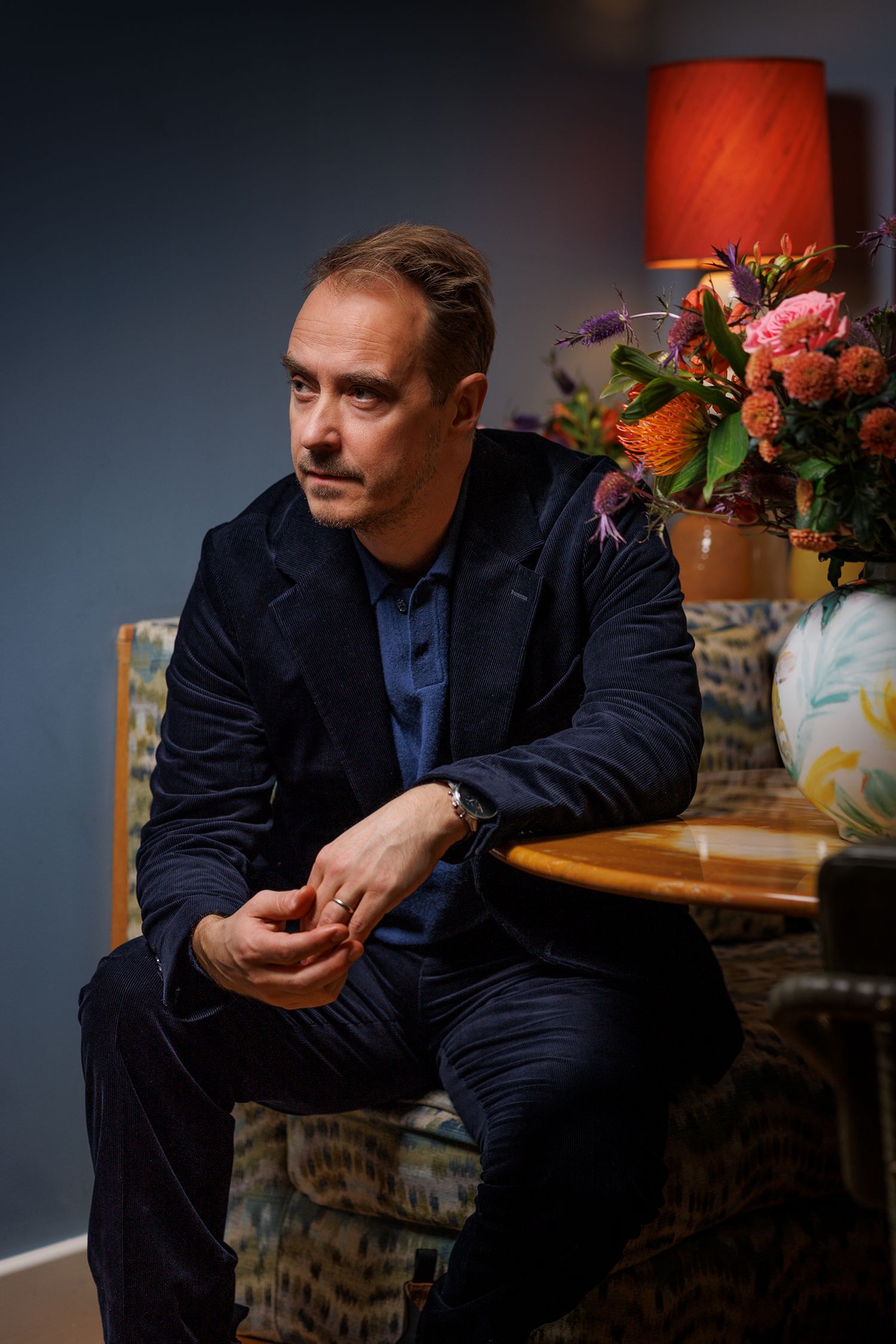 Person i mörk kostym sitter fundersamt nära blomsterarrangemang i stämningsfullt ljus