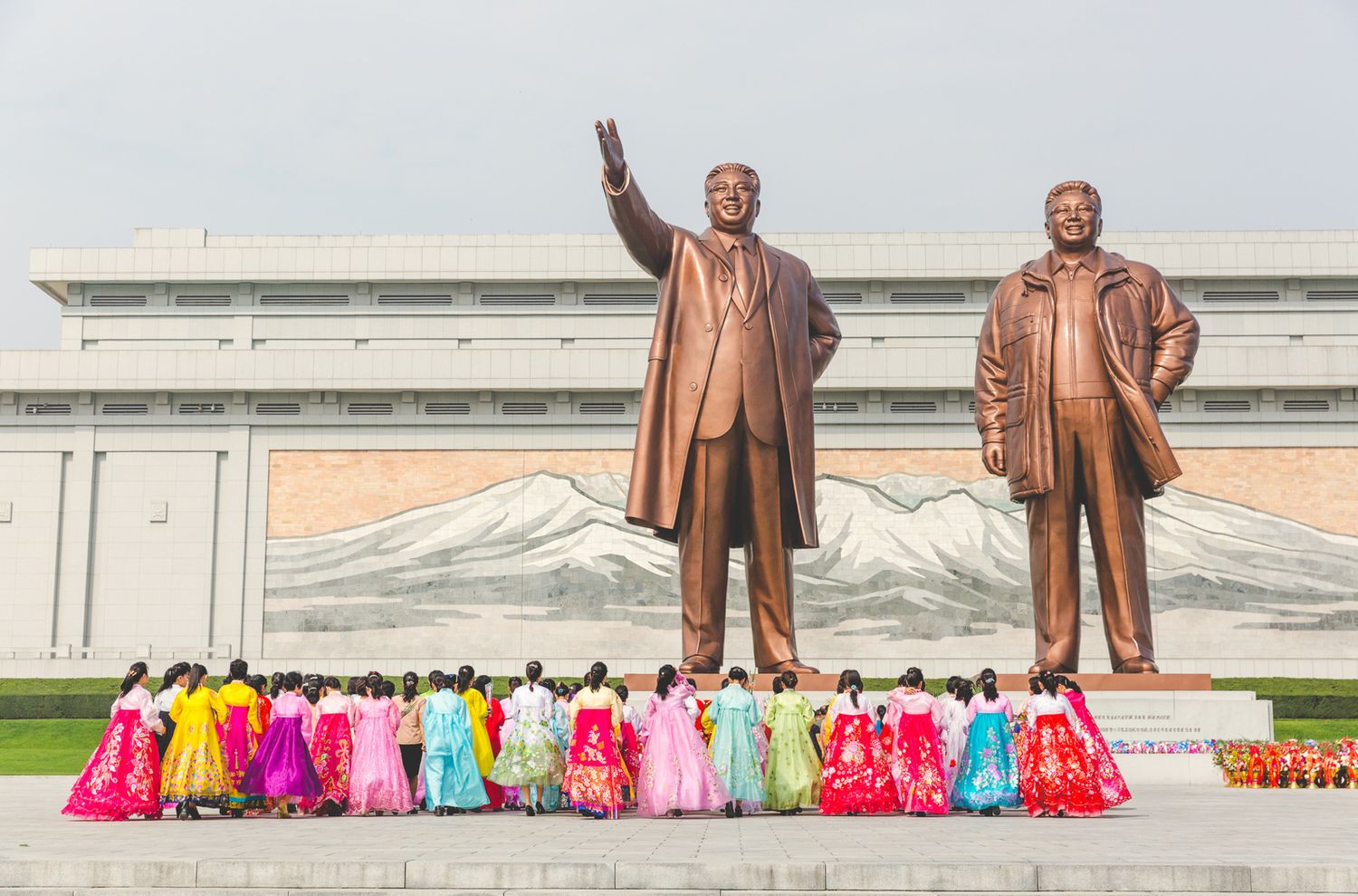 Colorfully dressed people arranged in rows before large bronze statues.