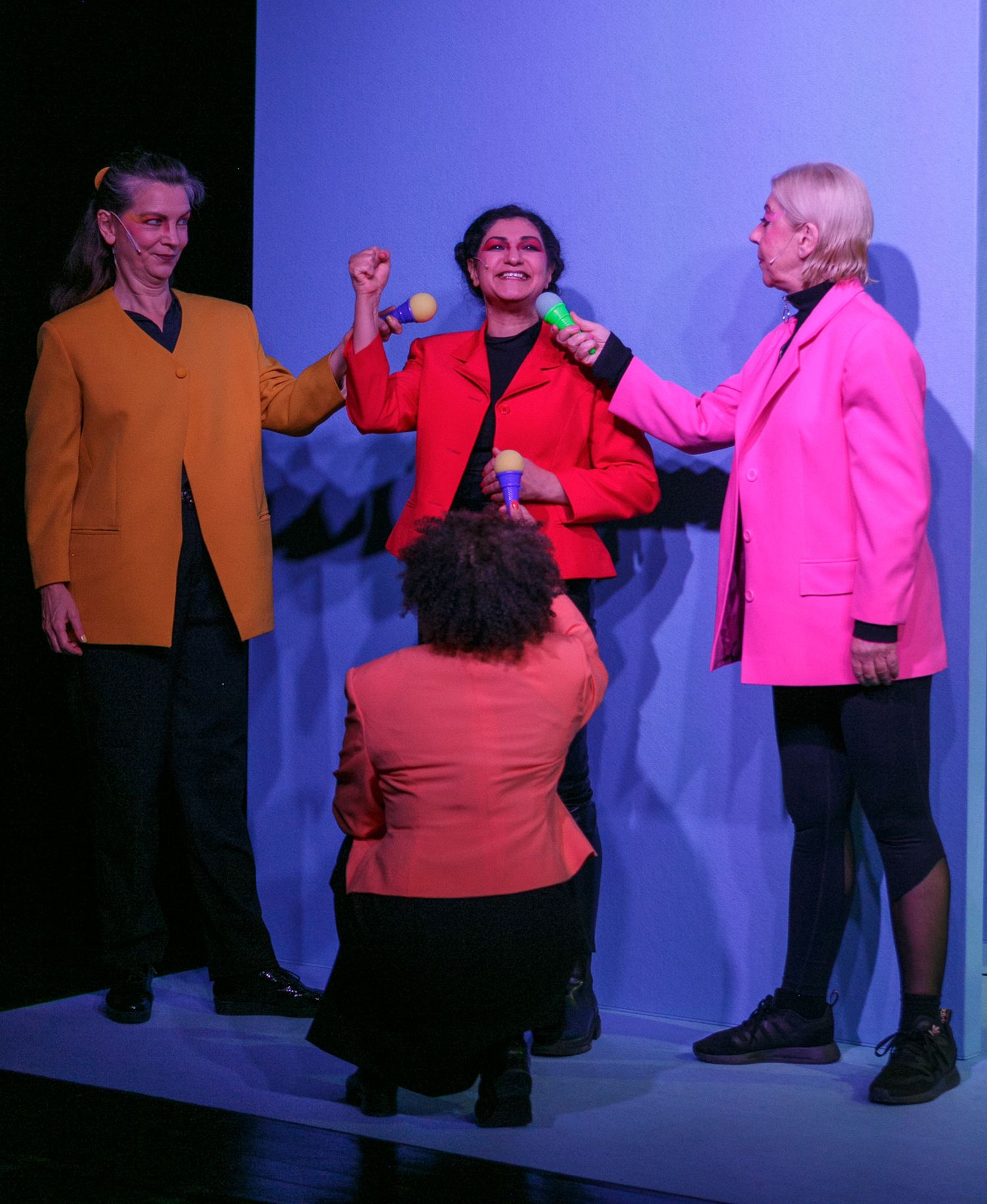 Theater actors in bright pink and orange costumes performing on a blue-lit stage.