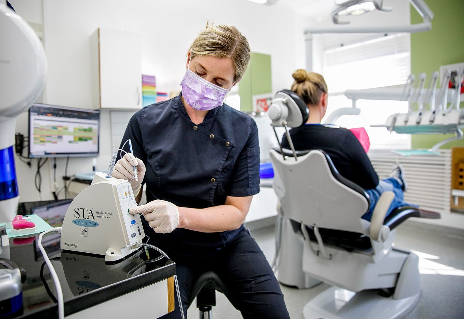 Dental professional in black scrubs and face mask working with equipment in modern clinic setting.