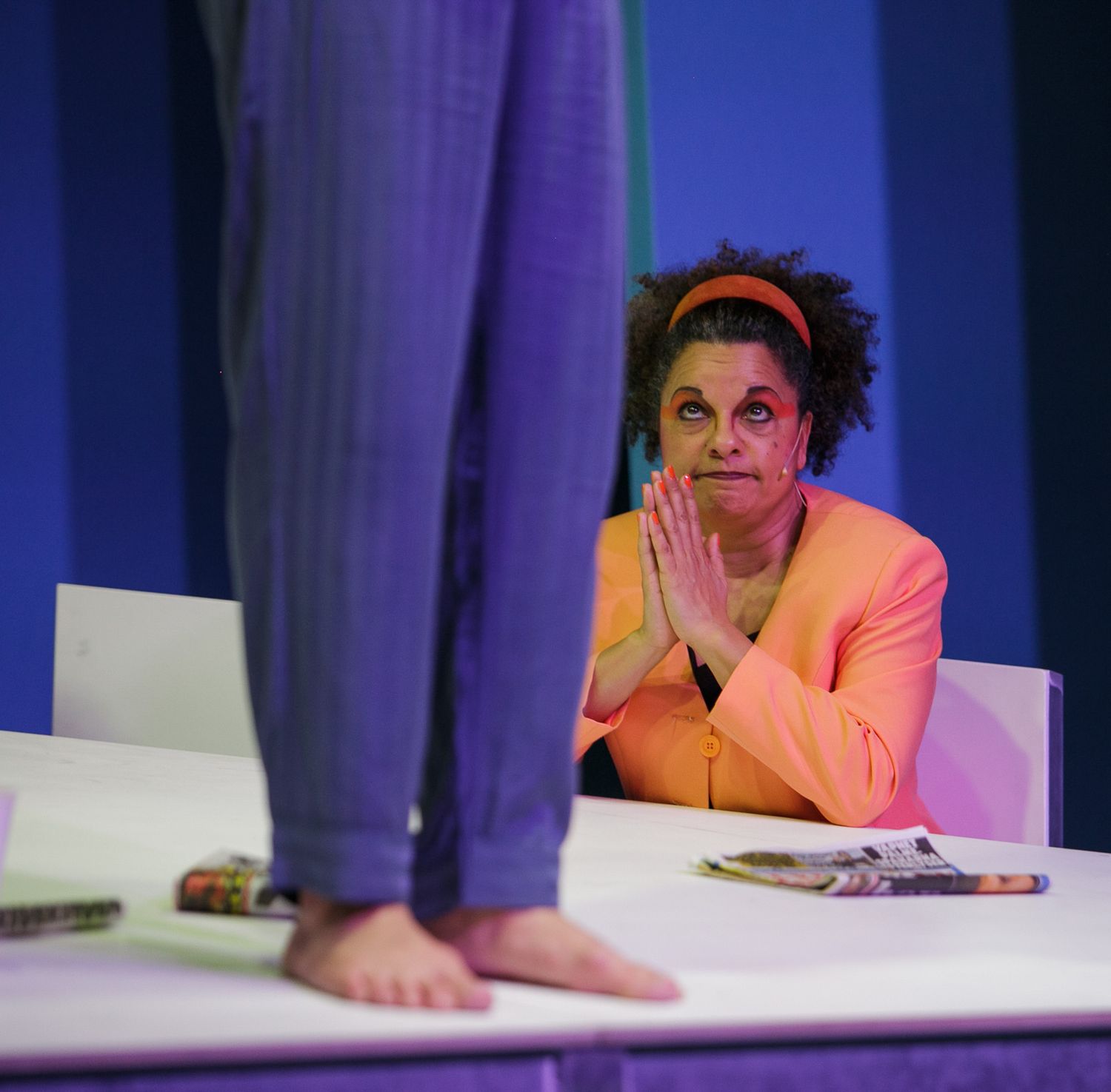 Stage performers in colorful outfits seated at a table during a theatrical production with blue lighting.