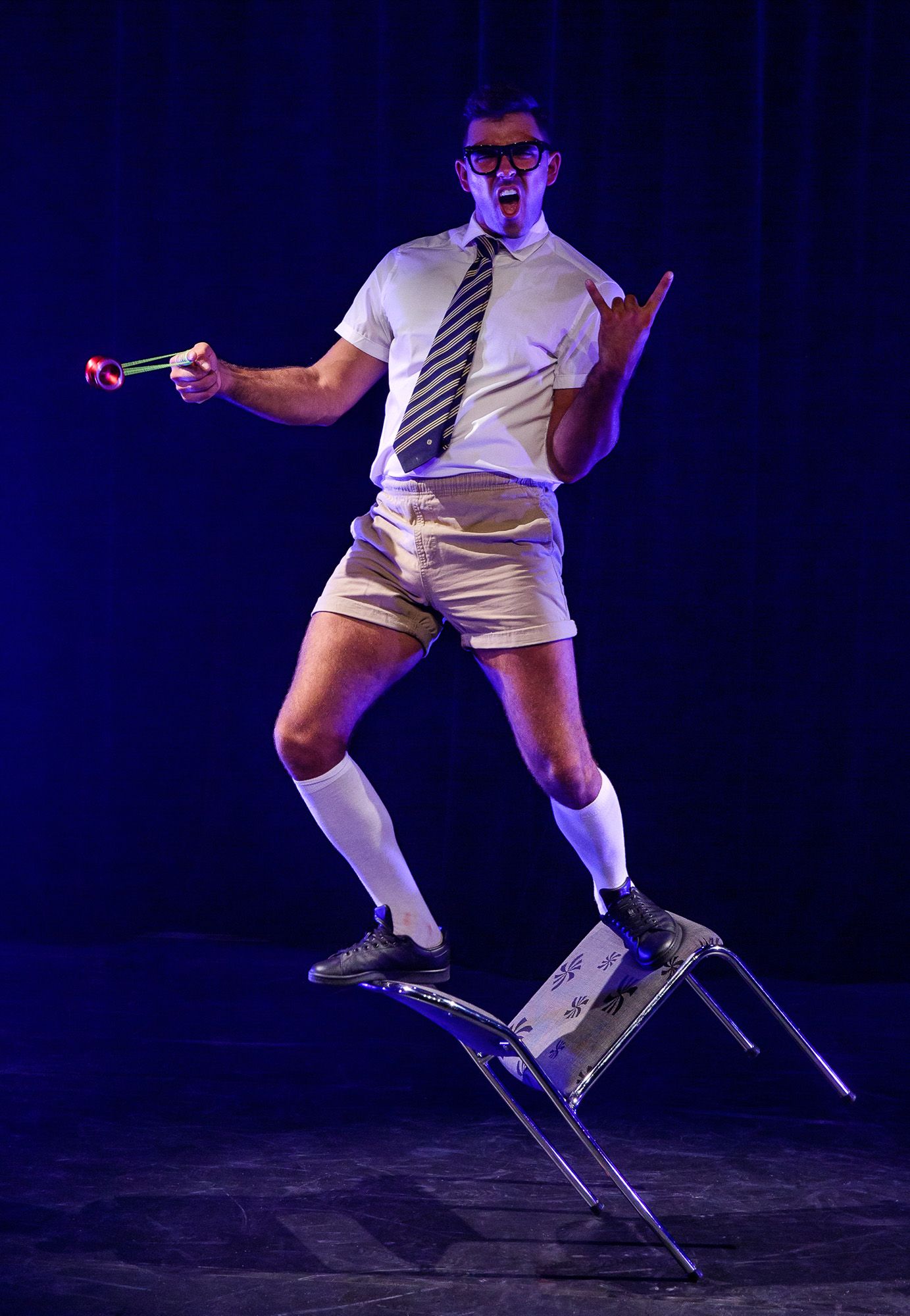 Performer in white outfit and tie demonstrates dynamic jumping movements on dark stage.