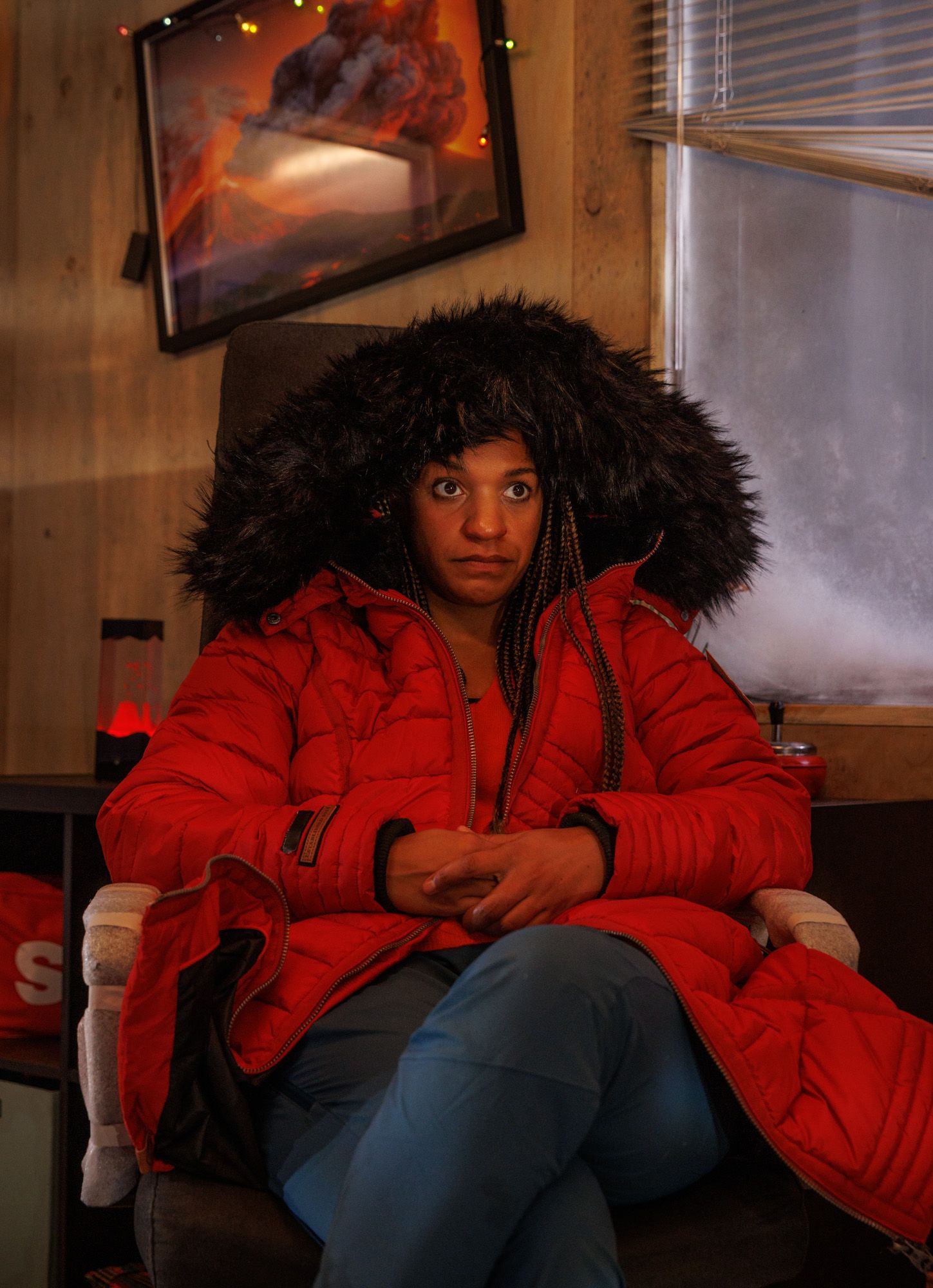 Person in red winter coat with fur hood sits in moody lighting.