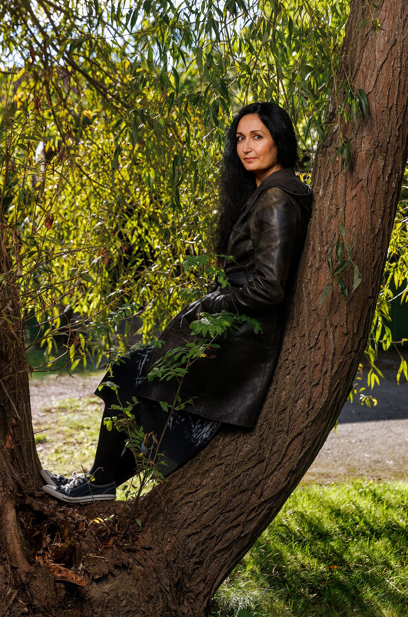 Figure in black leather jacket sitting against a tree trunk with green foliage in background.