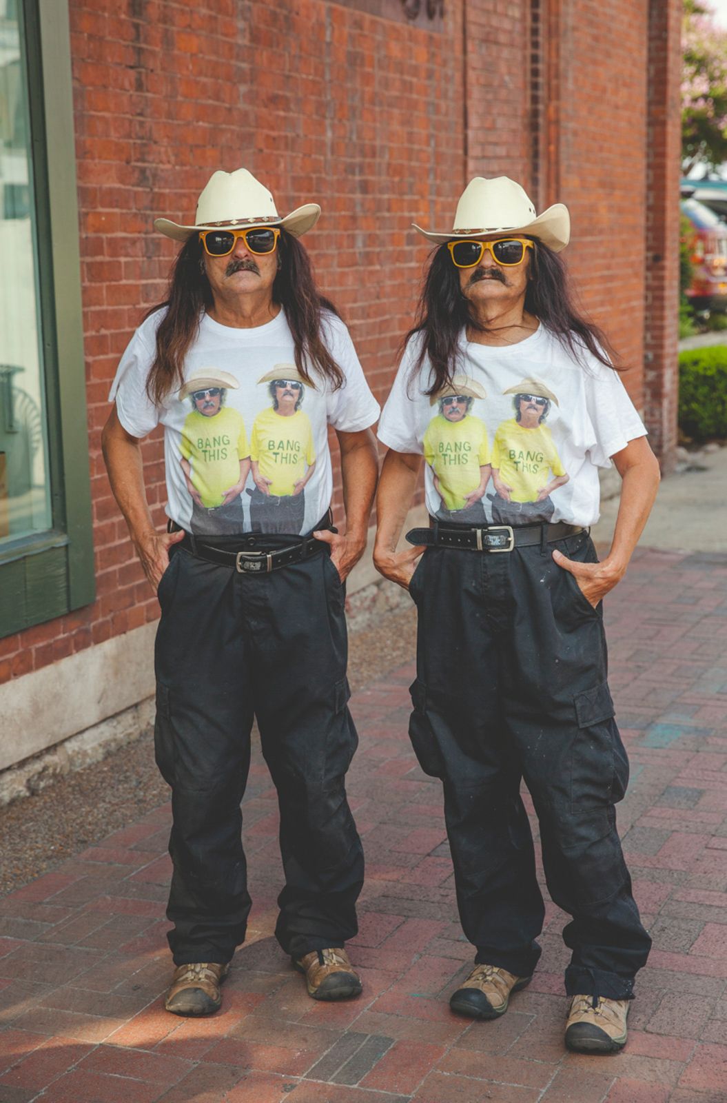 Two people in matching outfits and cowboy hats pose against a brick wall background.