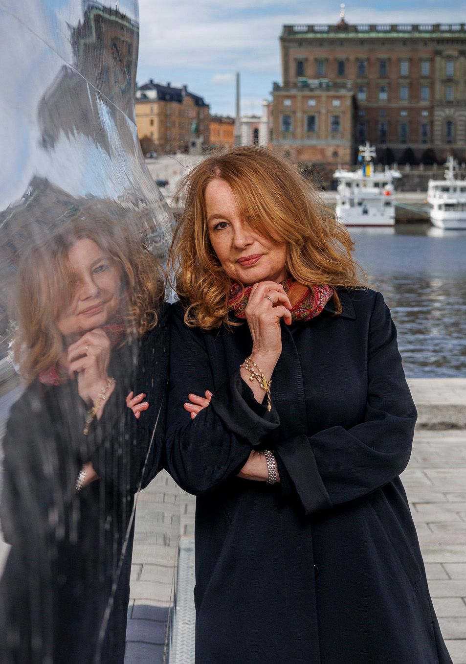 A woman in a black coat poses by the waterfront with historic buildings and boats in the background.