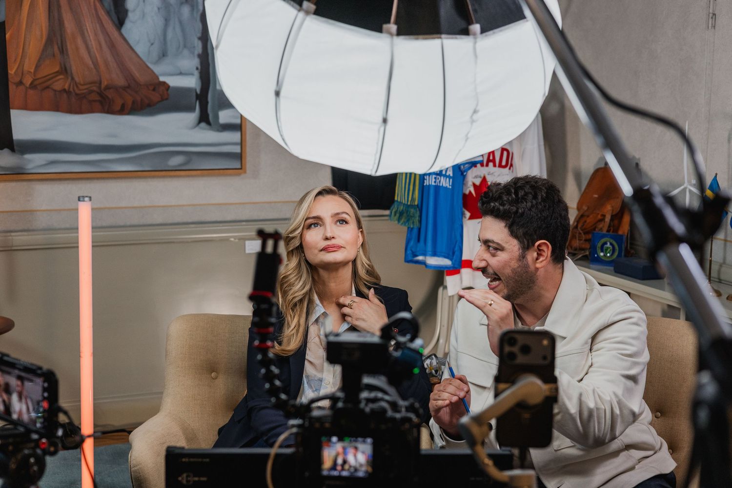 Two people sit together in front of ring light and cameras during a professional video or photo shoot indoors.