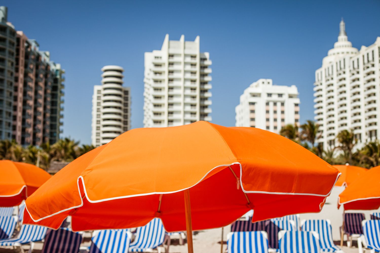 Vibrant orange and blue striped beach umbrellas line a sunny beachfront with high-rise buildings in the background.