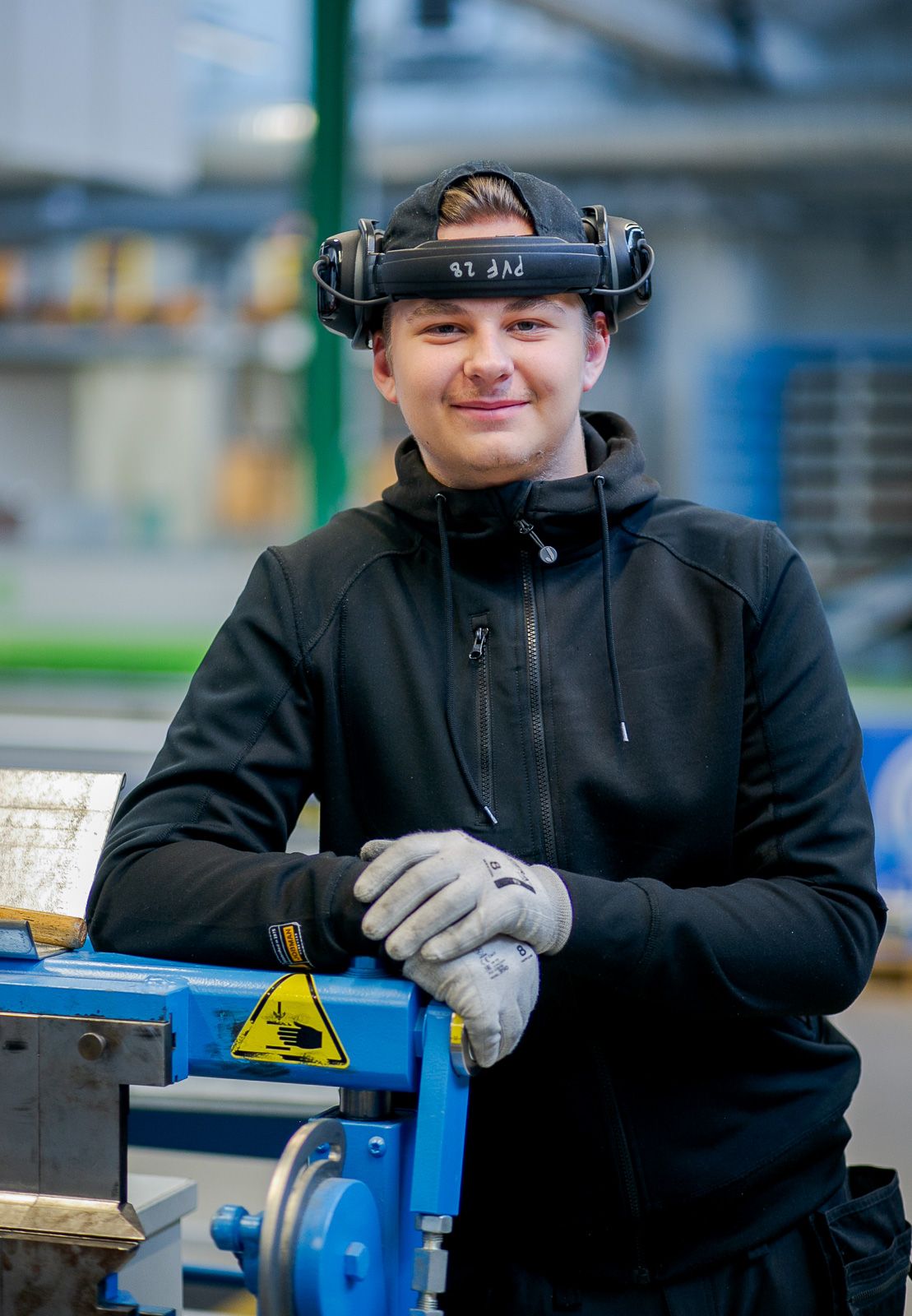 Worker in black hoodie operating blue industrial equipment in workshop.
