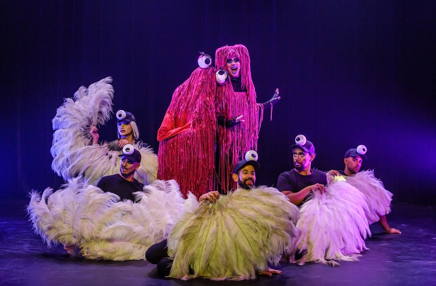 Theatrical performance featuring dancers in pink and yellow costumes on dark stage.
