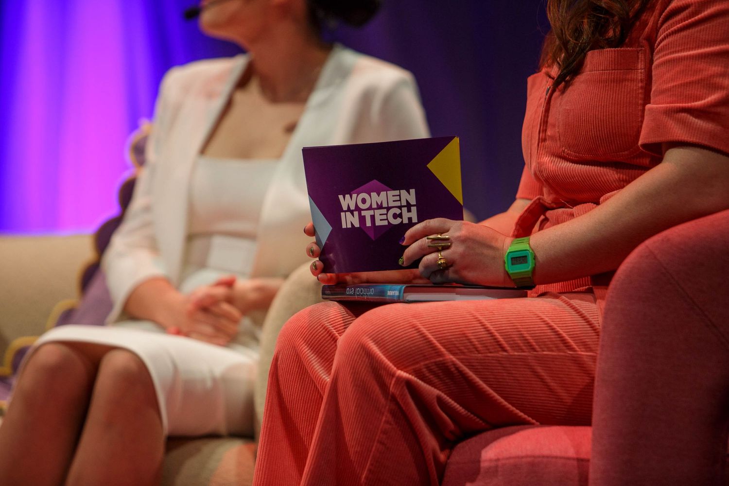 Attendees seated in red chairs during a conference session with purple ambient lighting.