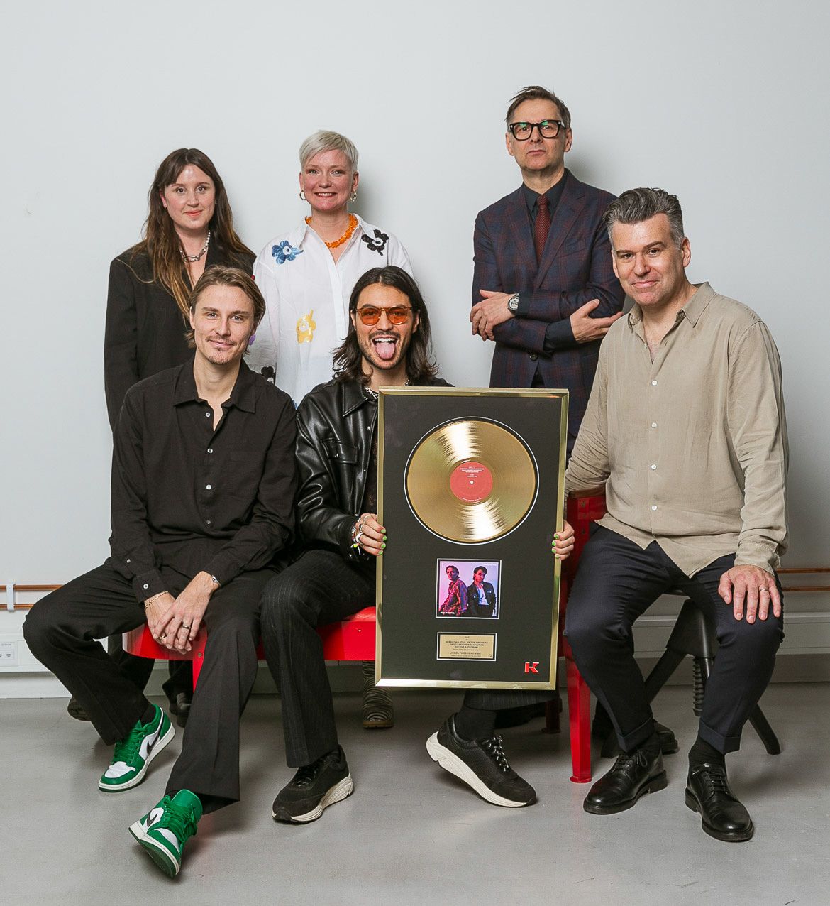 People seated together displaying music industry achievement awards against white backdrop.