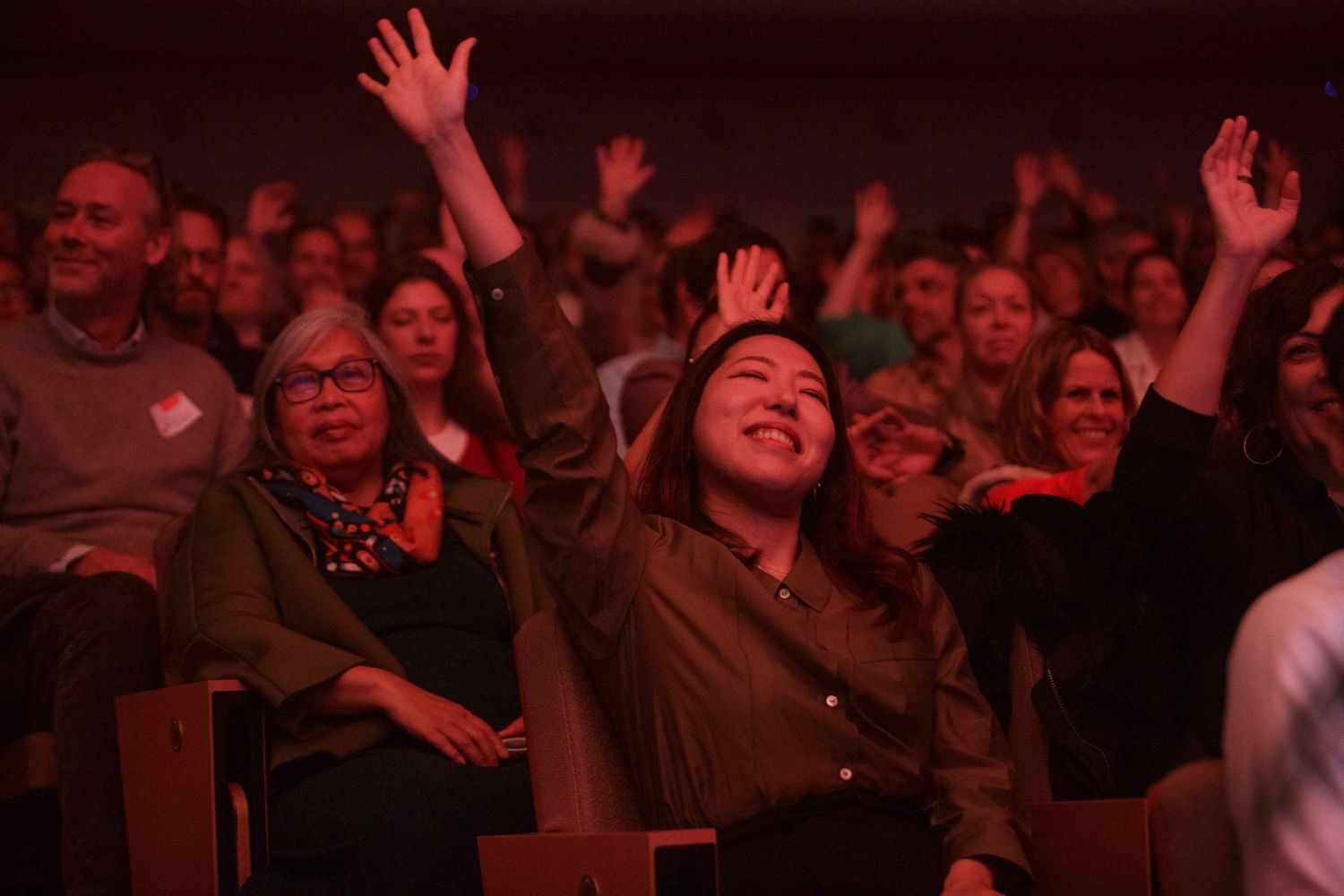 An enthusiastic audience with raised hands illuminated in red lighting at an event.