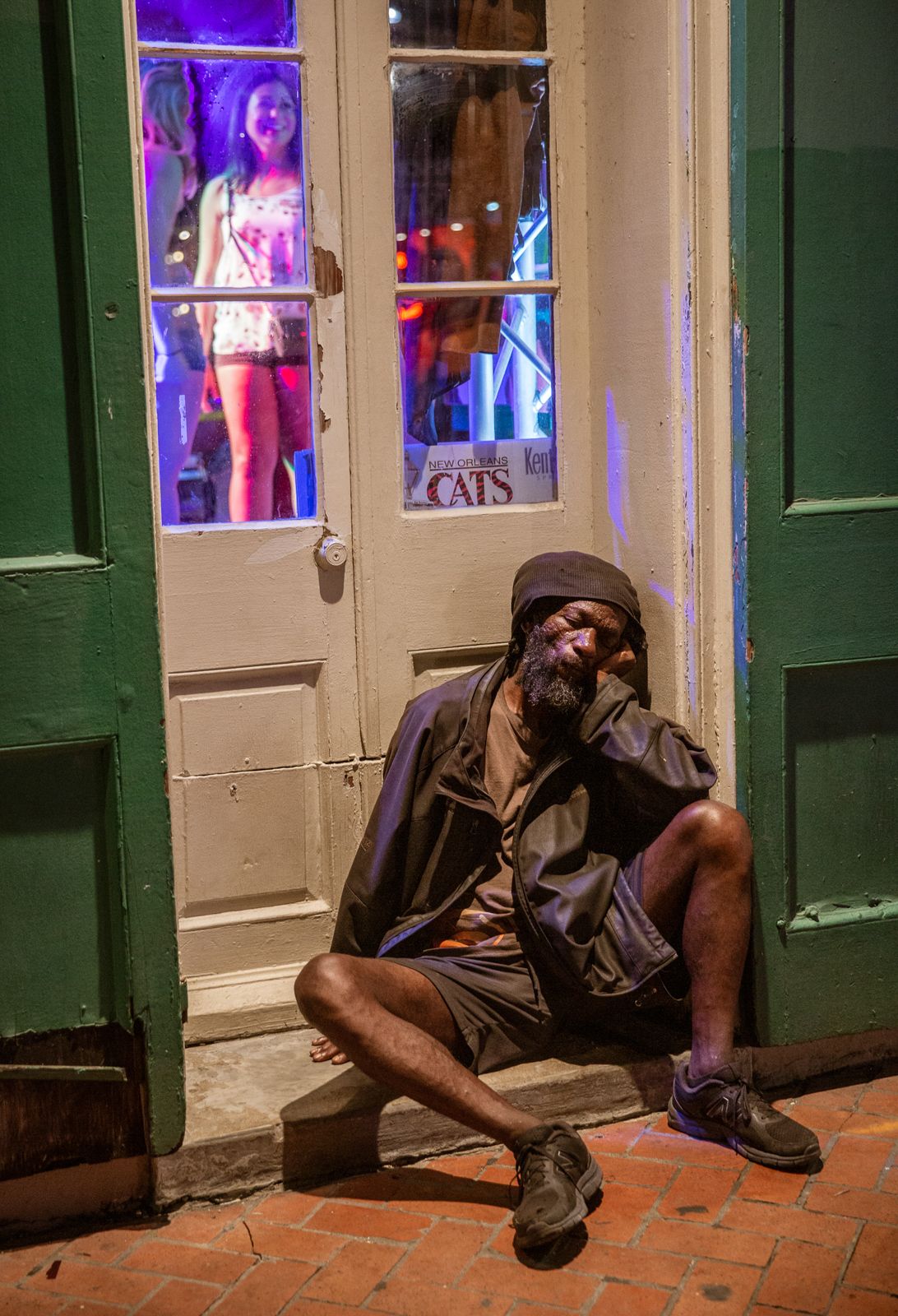 Person sleeping on sidewalk outside illuminated storefront with purple neon lights.